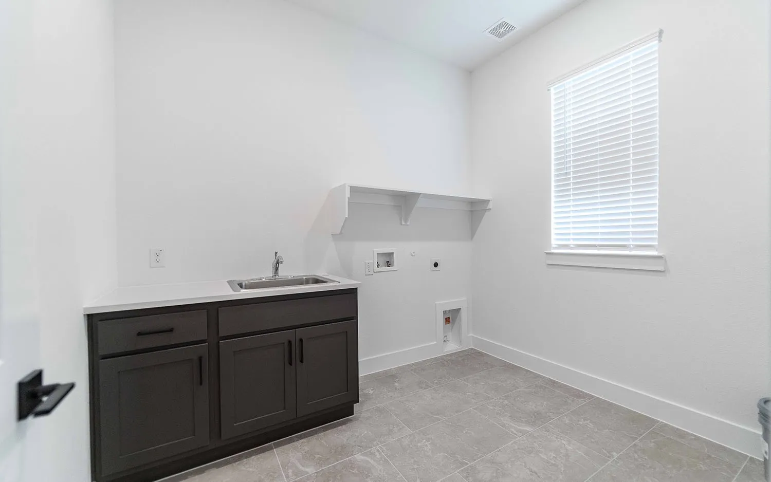 Utility space featuring a window with blinds, a built-in sink with a base cabinet, a white countertop, two wall-mounted shelves, and tile flooring