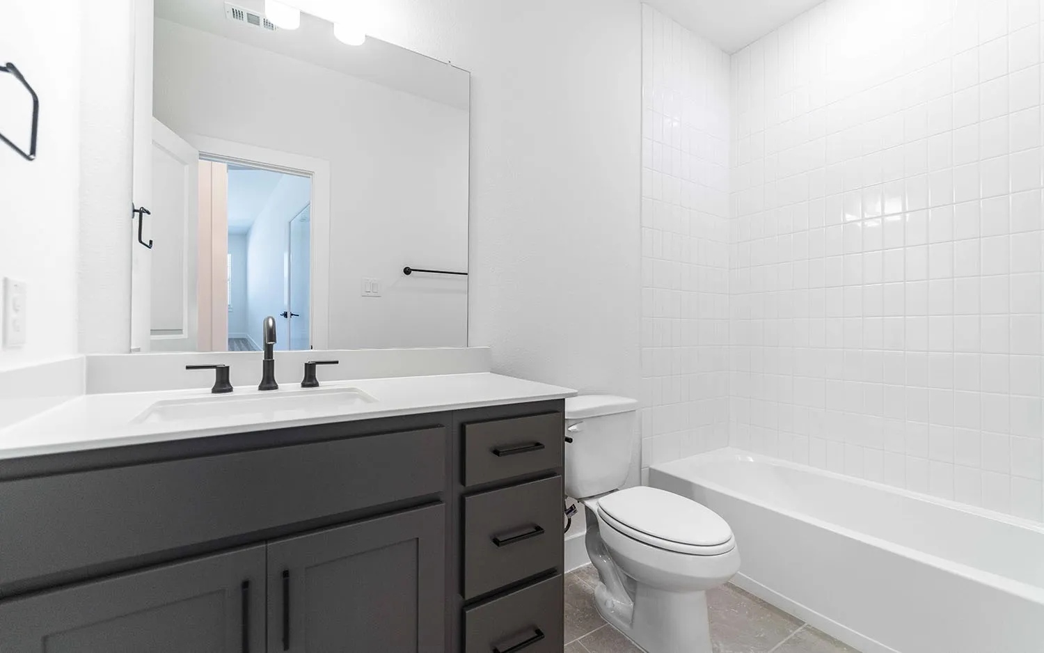Bathroom featuring a white subway tile tub surround, a dark grey vanity with a white countertop and integrated sink, a matte black faucet, and white walls