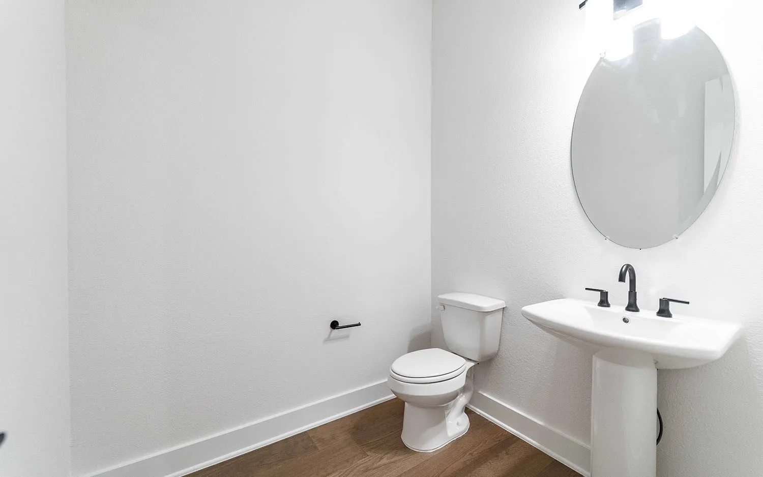 Pedestal sink with matte black hardware, oval mirror, white toilet, wood-finish flooring, and white baseboards