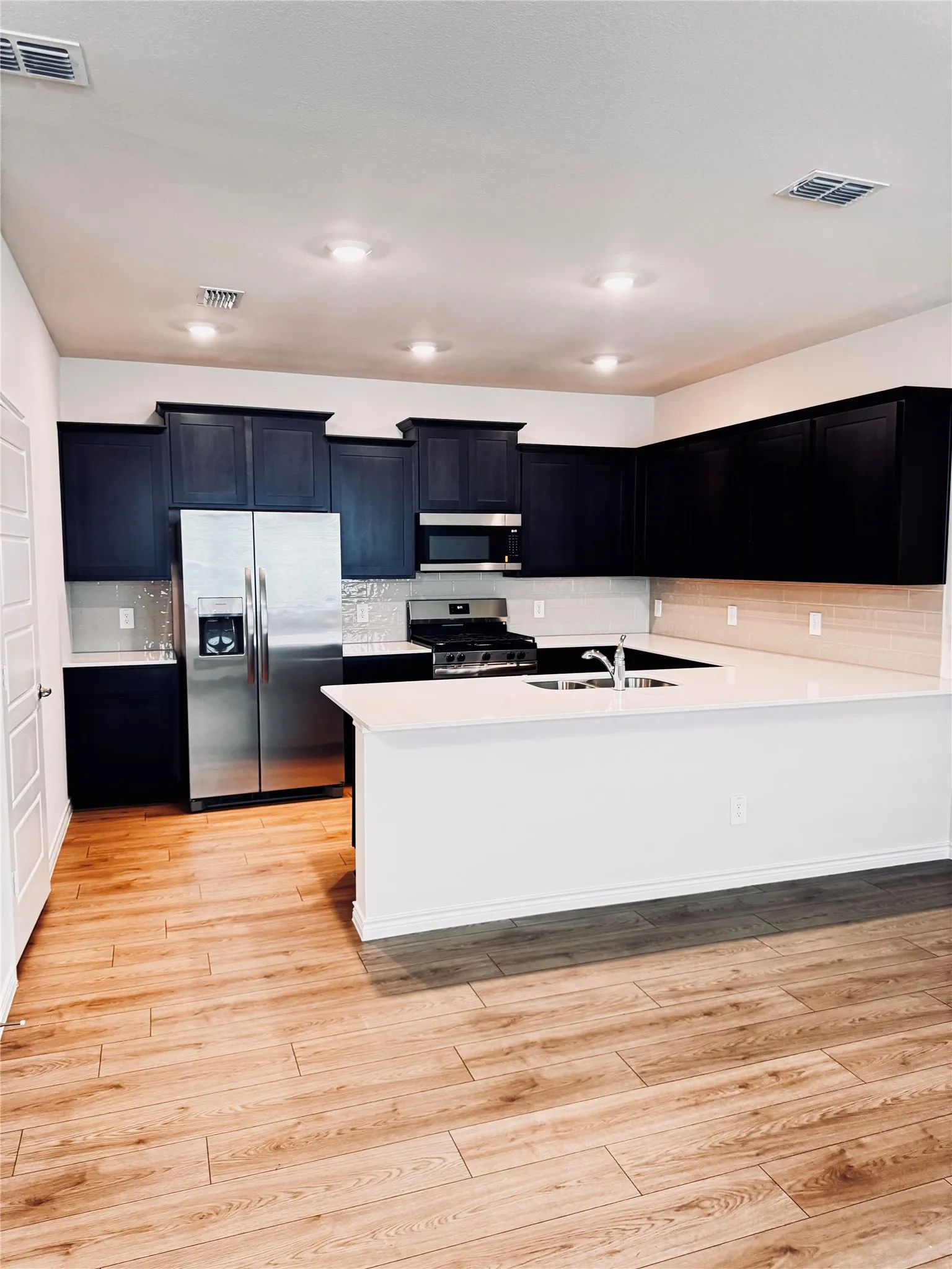 Modern kitchen featuring dark cabinetry, stainless steel appliances, a center island with sink, light countertops, and wood-finish flooring