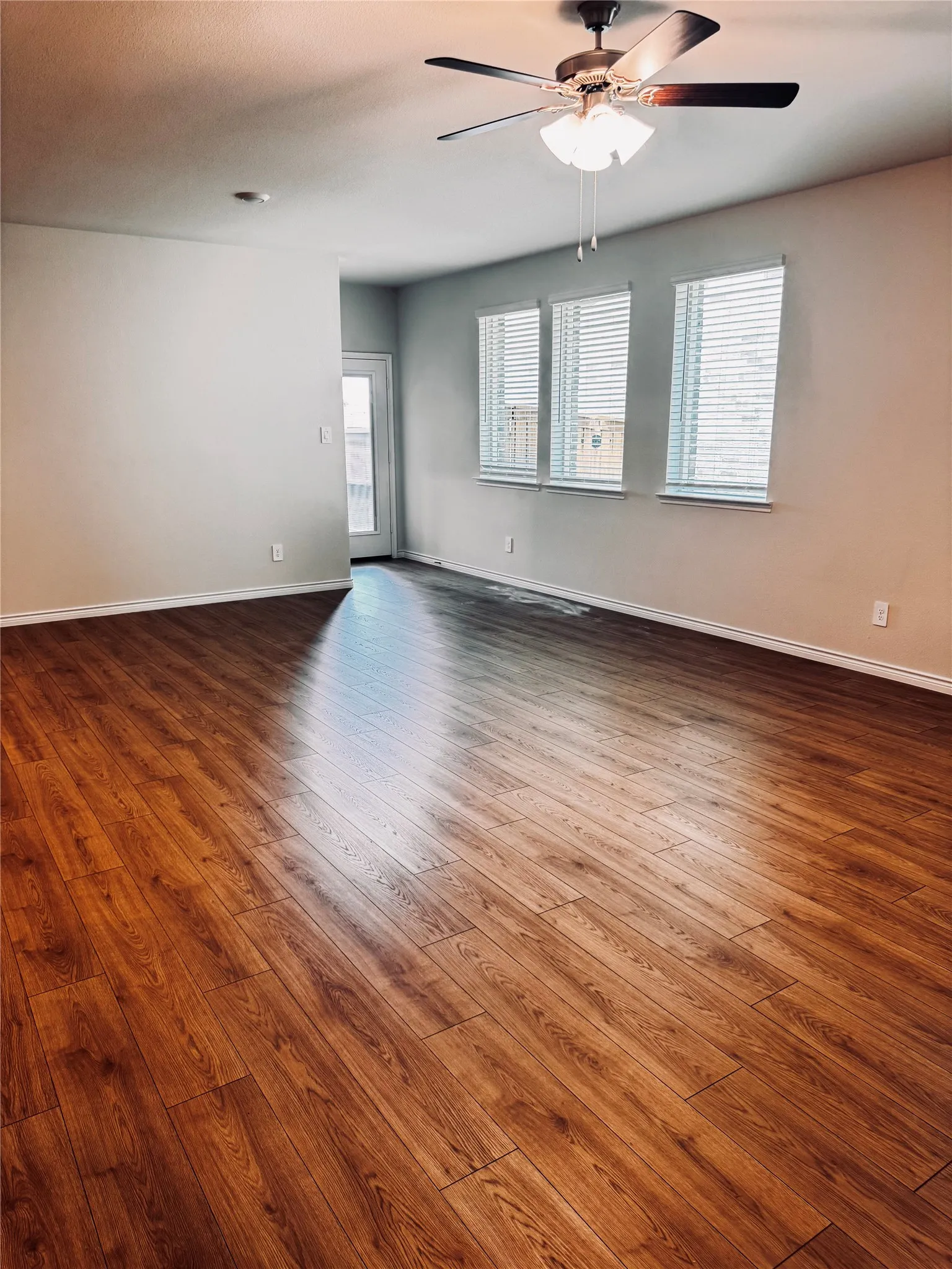 Spacious living room featuring wood-finish flooring, a ceiling fan with integrated lighting, multiple windows with blinds, and a glass-paneled door