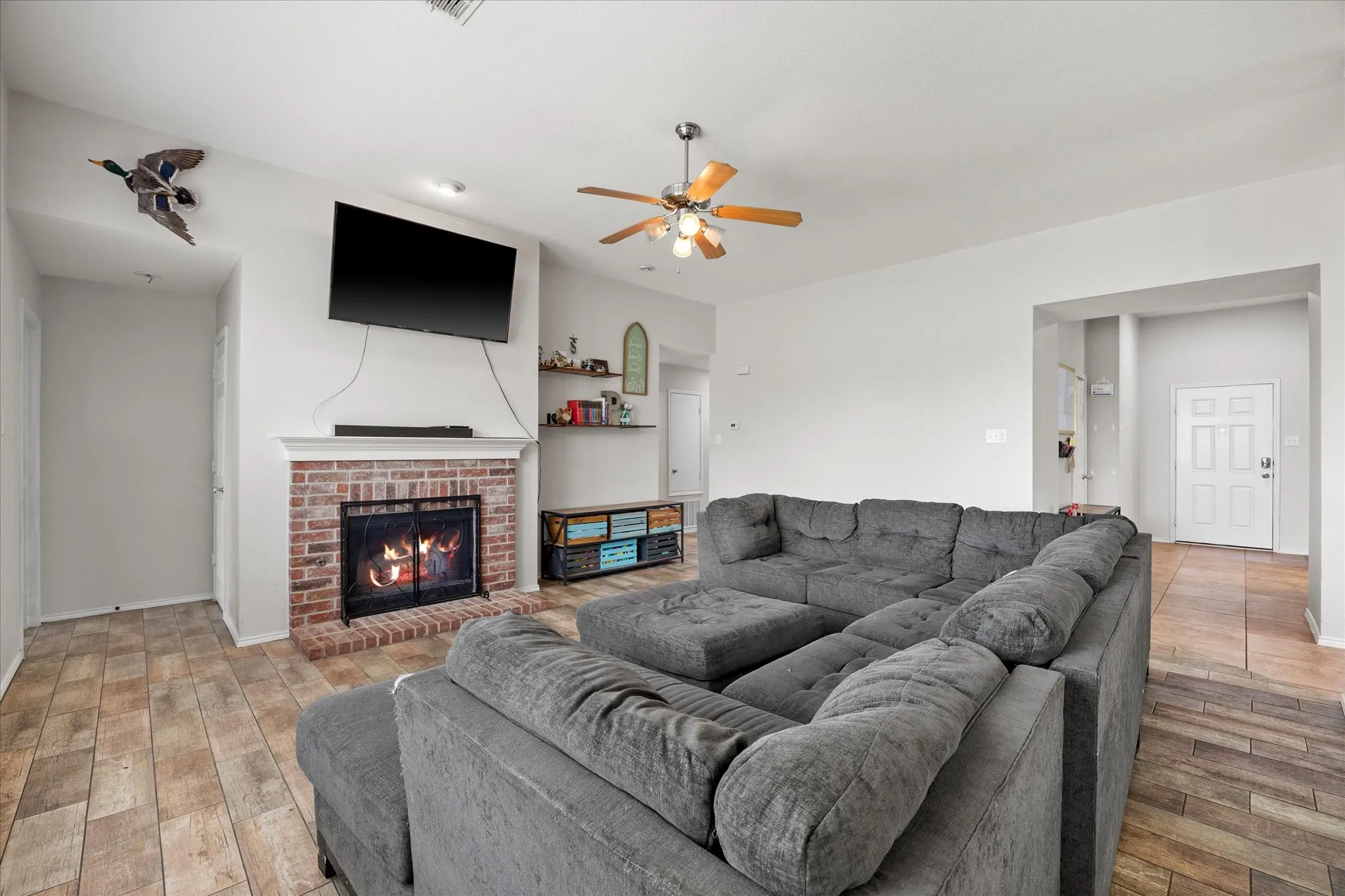Spacious living area featuring a brick-surround fireplace with white mantel, a ceiling fan with integrated lighting, and wood-finish flooring