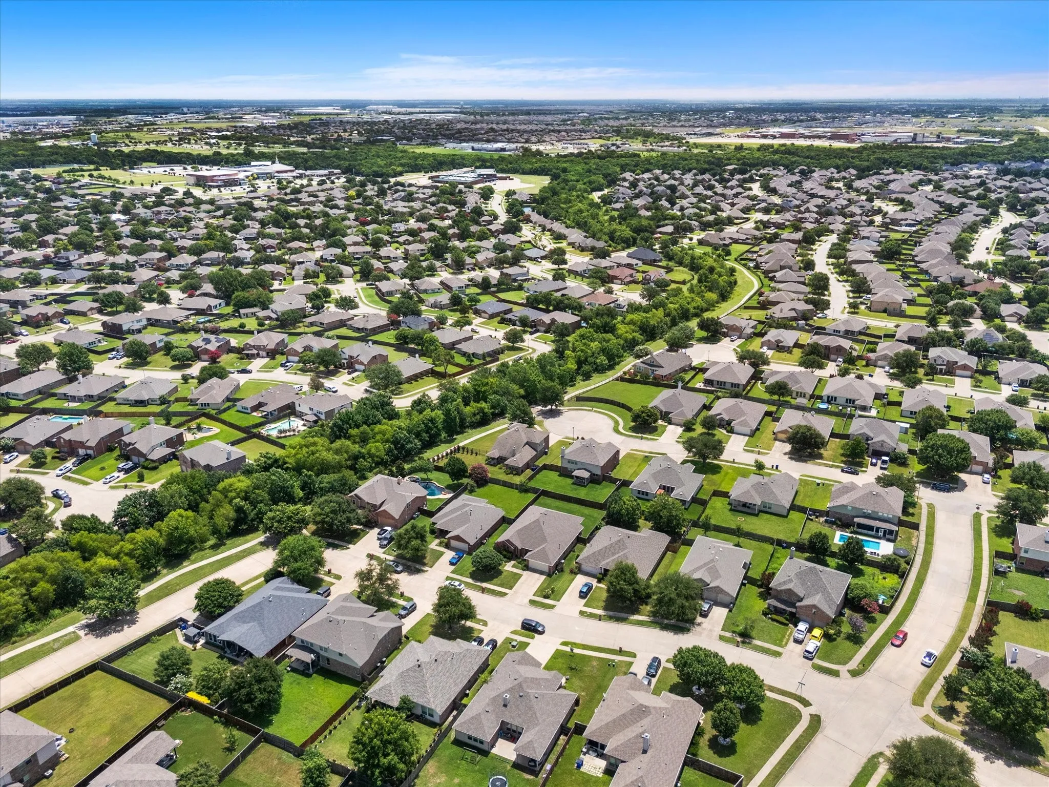 Aerial perspective showcasing a residential community with winding streets, mature landscaping, individual properties with green lawns, and a distant tree line