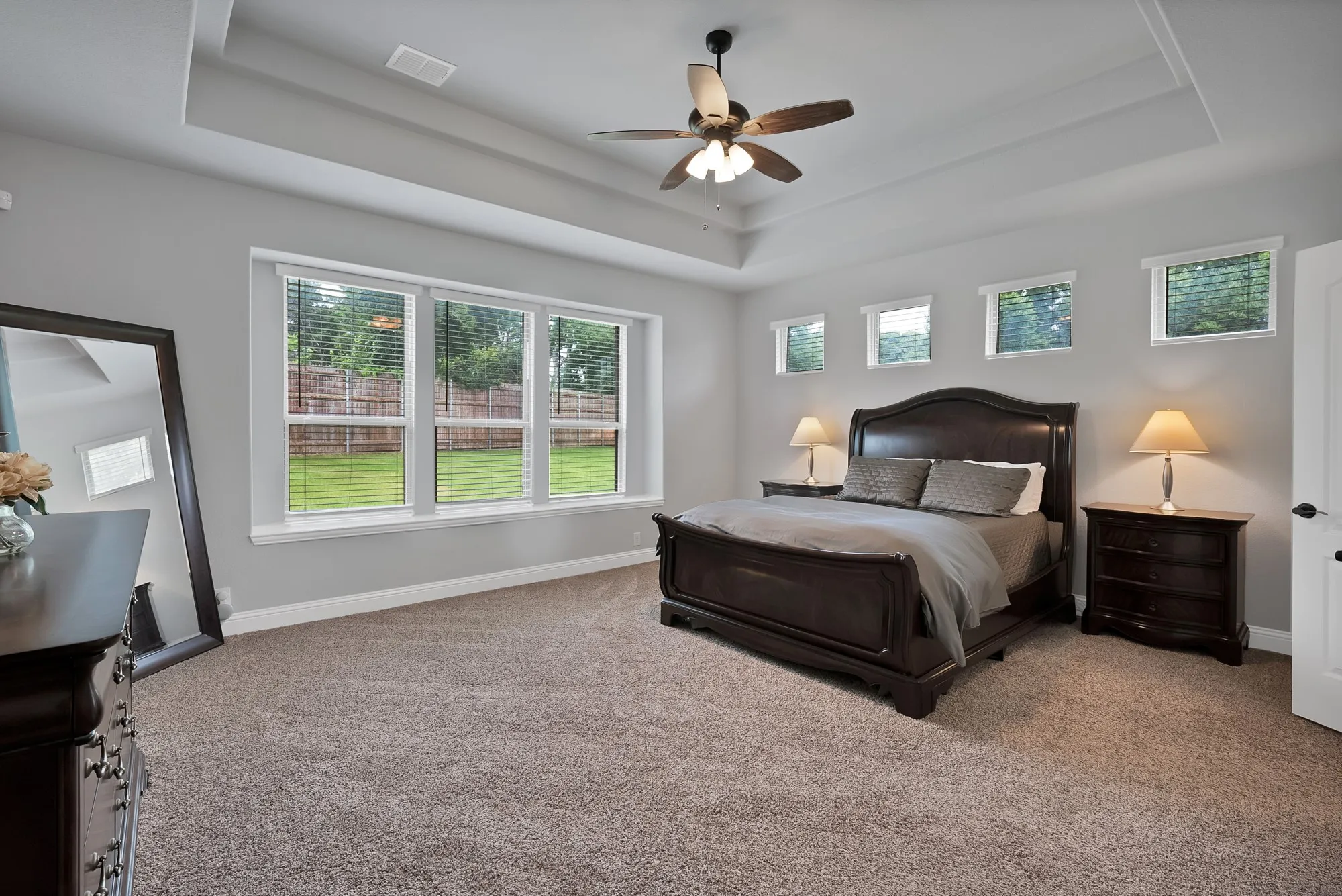 Spacious bedroom featuring a tray ceiling with a ceiling fan, plush carpet flooring, and a bank of three large windows providing ample natural light