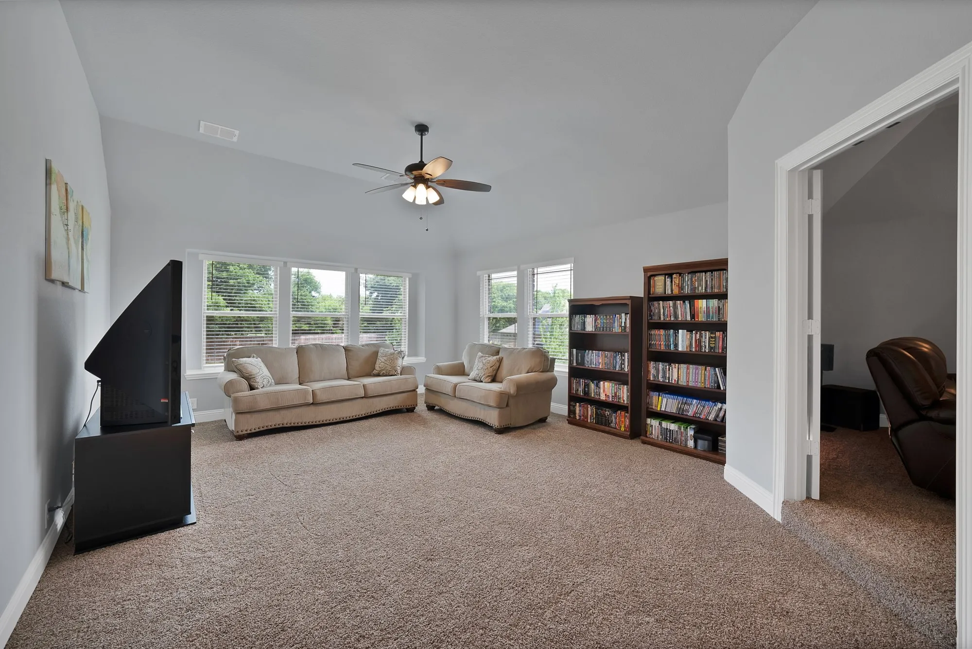Carpeted entertainment room featuring vaulted ceilings, multiple windows with blinds, and an overhead ceiling fan with lighting