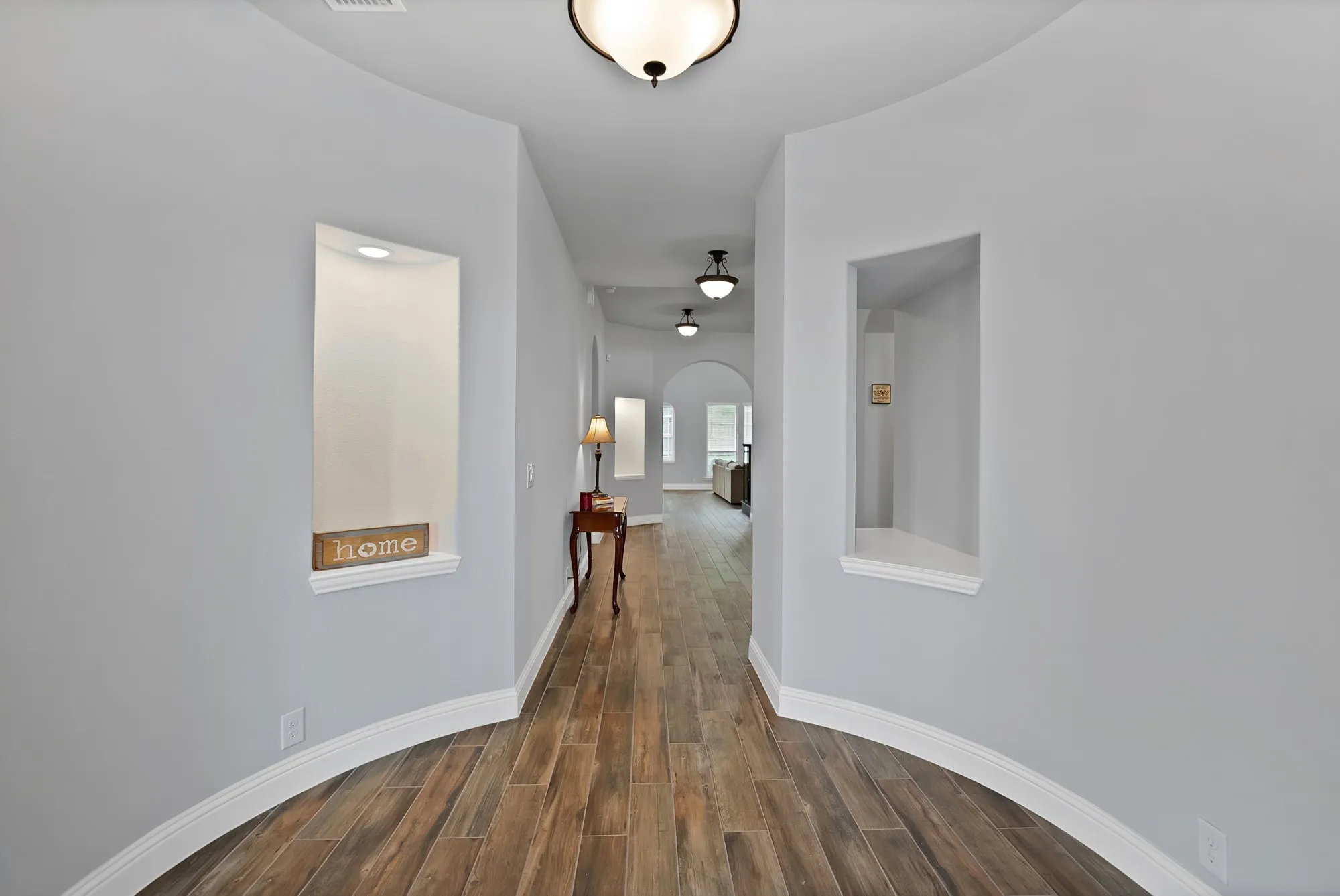 Hallway featuring wood-finish flooring, light gray walls, coved baseboards, recessed display niches, and ceiling-mounted light fixtures