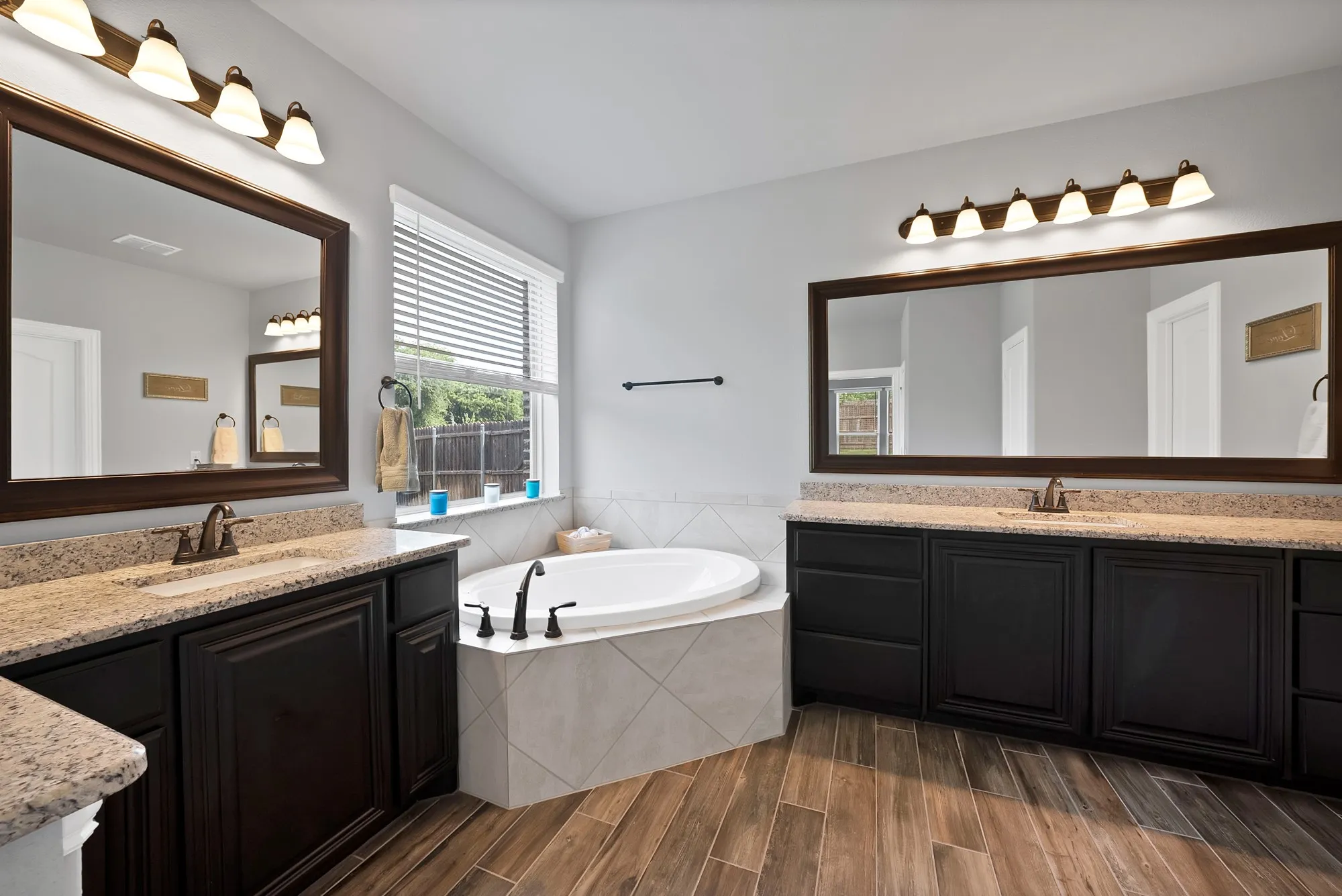 Spacious bathroom featuring a corner soaking tub, dual vanities with light-toned countertops, wood-finish flooring, and a large window with blinds
