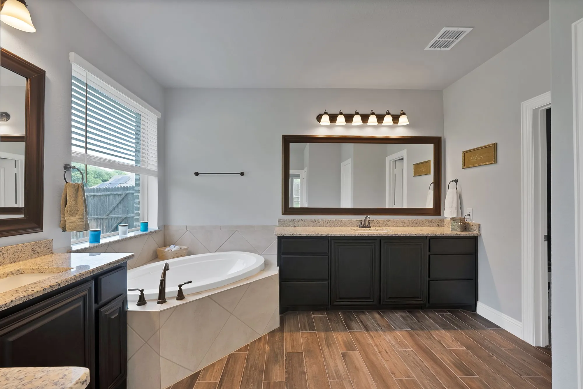 Soaking tub with tiled surround, dual vanities featuring granite countertops and dark wood cabinetry, wood-finish flooring, and a large window with blinds