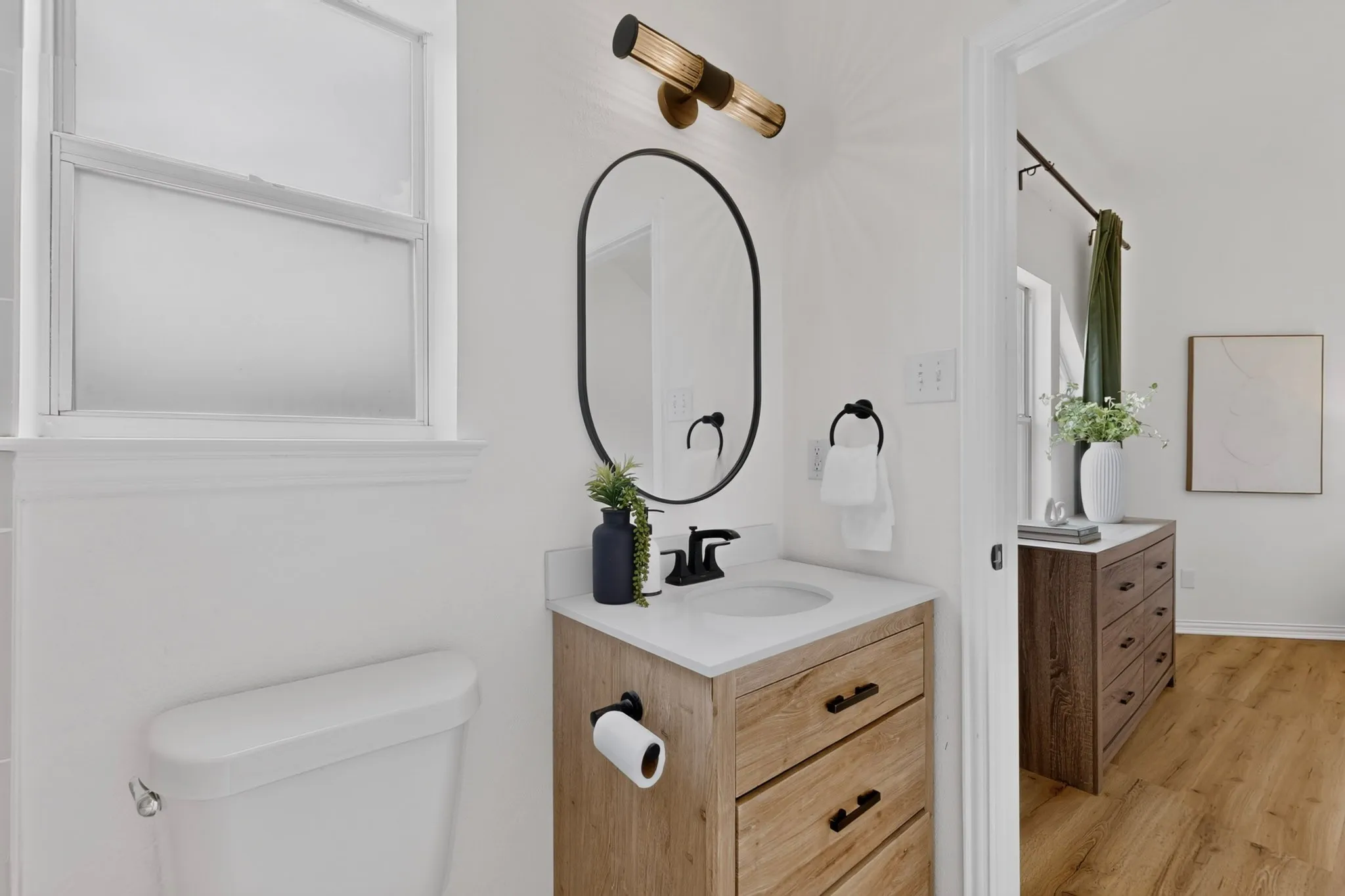 Bathroom vanity featuring a light wood-finish cabinet with black hardware, a white countertop, and an oval-framed mirror
