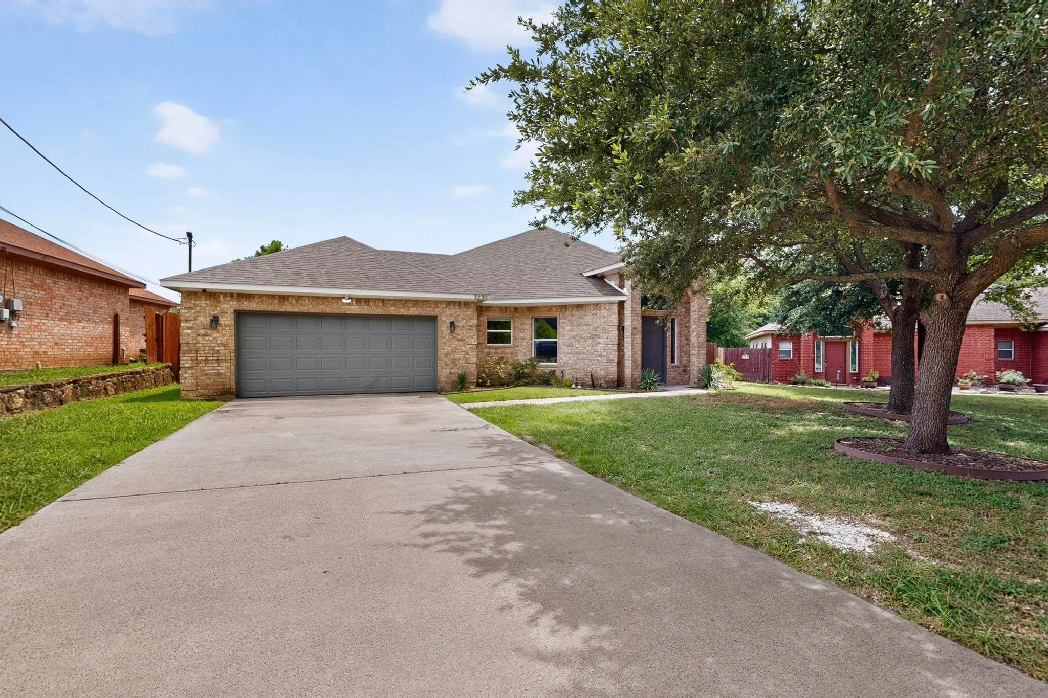 Brick exterior featuring a two-car garage with a gray door, an extended concrete driveway, and mature trees framing the front lawn