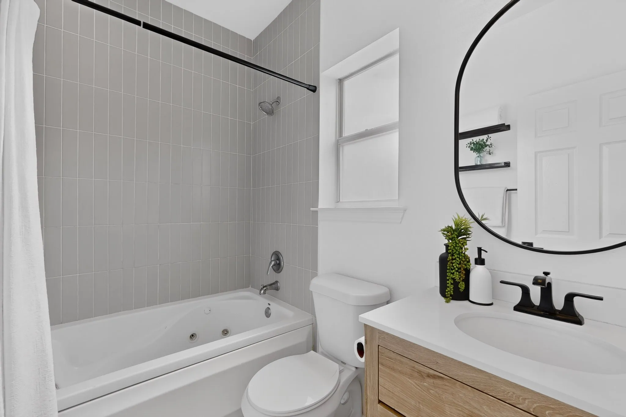 Bathroom featuring a jetted bathtub with subway tile surround, a single window, an oval mirror, a wood-finish vanity with an integrated sink, and black fixtures