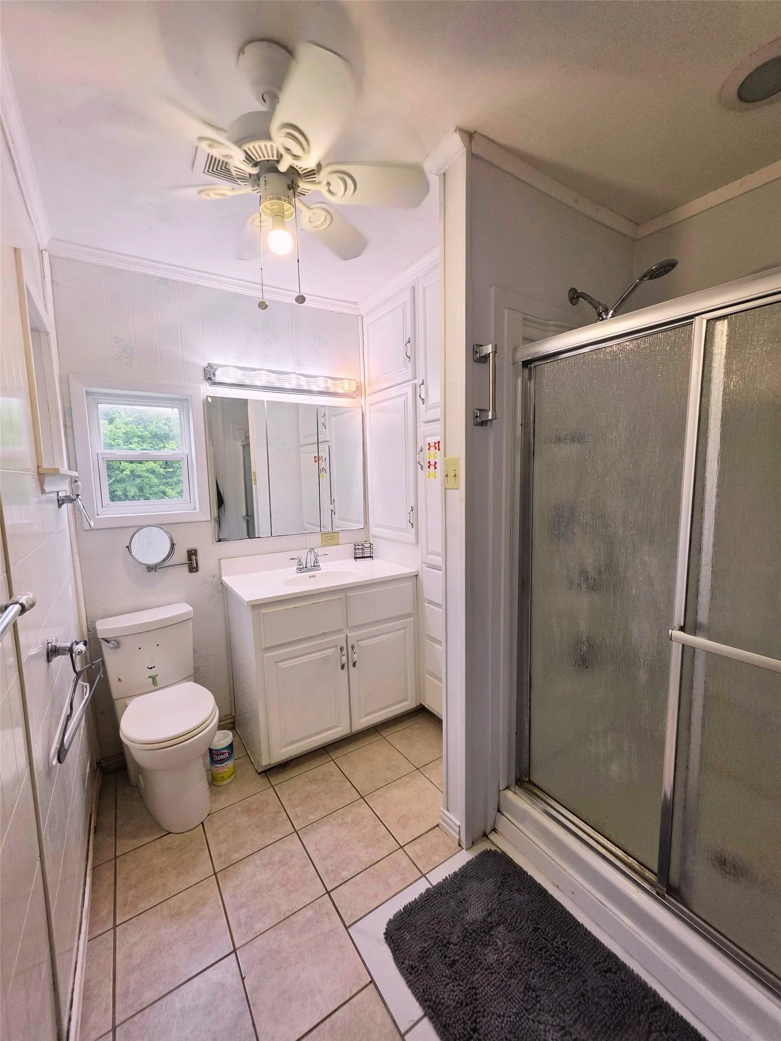 Bathroom featuring a white vanity with integrated sink, a glass-enclosed shower, tiled flooring, a window, and a ceiling fan