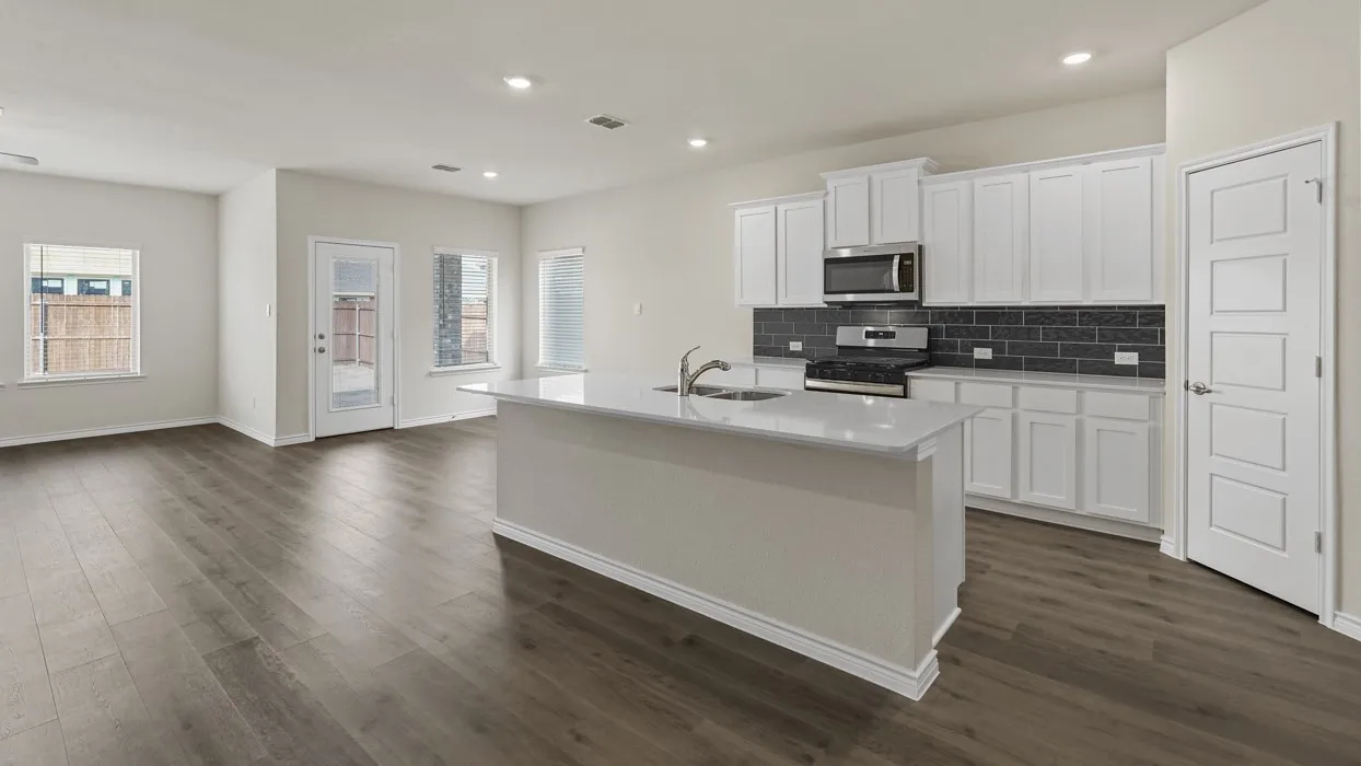 Open-concept living space featuring wood-finish flooring, a kitchen island with an integrated sink, white cabinetry, a stainless steel microwave and range, and a dark subway tile backsplash