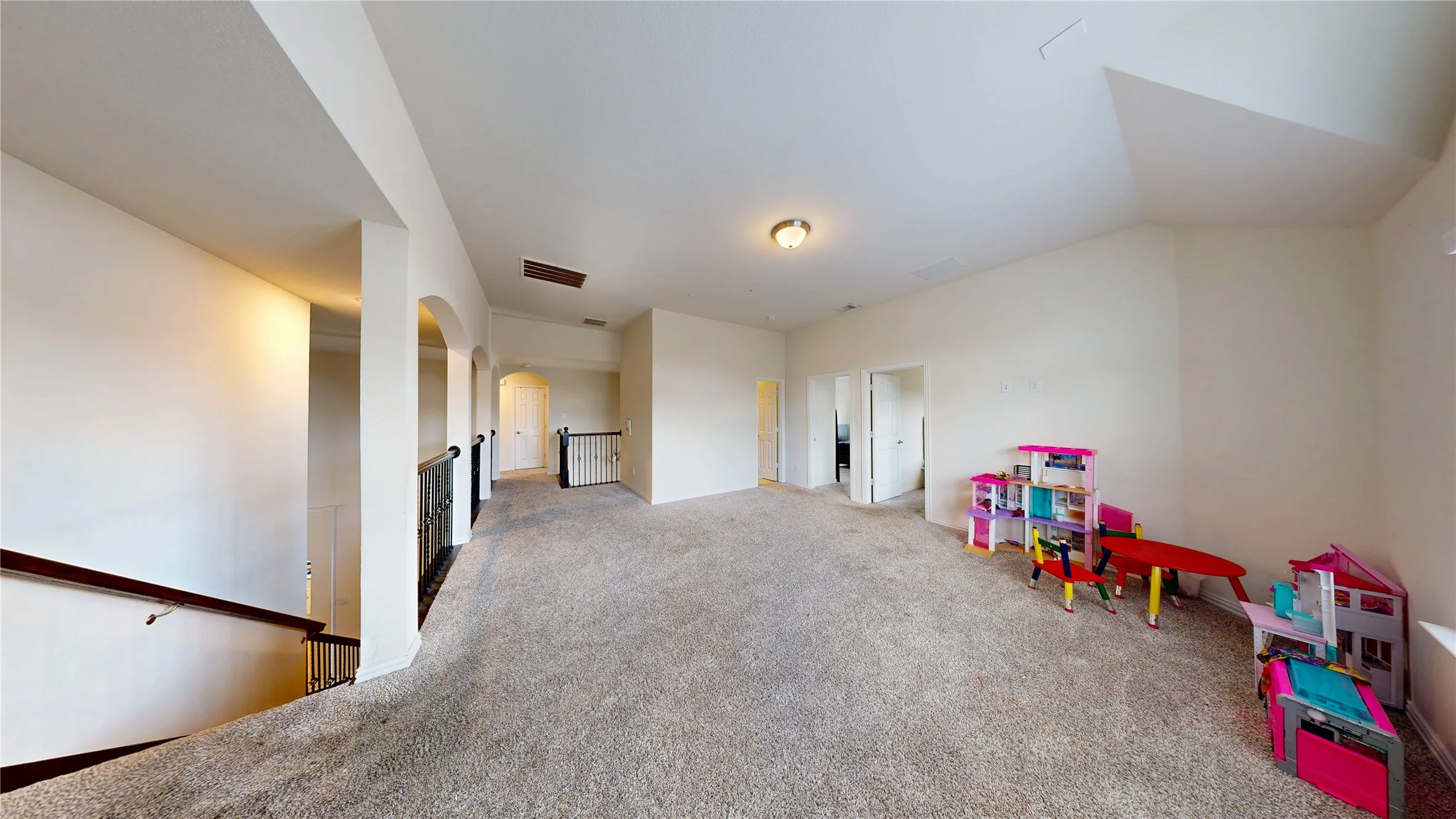 Spacious carpeted open area featuring arched doorways, staircase with dark wood handrail and wrought iron balusters, and recessed ceiling lighting