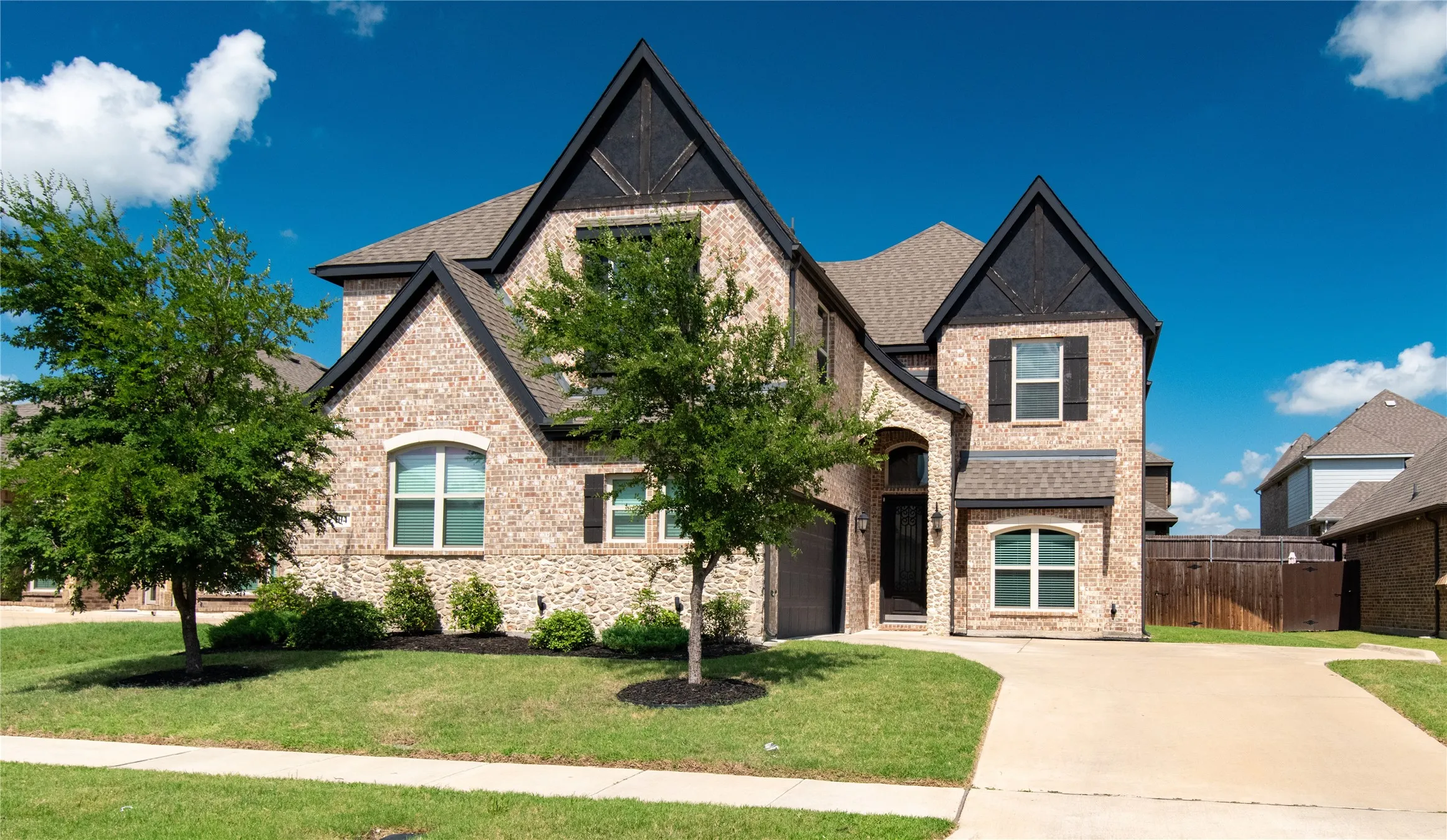 Brick and stone facade residence featuring a peaked roofline, dark trim accents, shuttered windows, an arched entry, and an integrated garage
