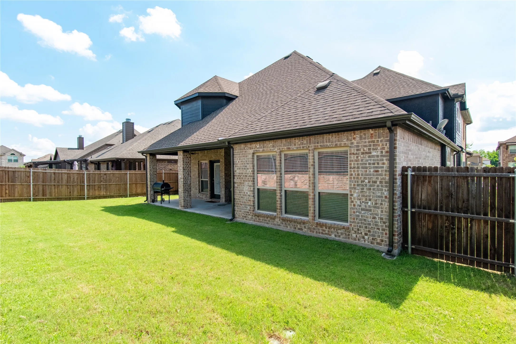 Expansive green lawn with a covered concrete patio, brick exterior, dark brown fence, and multiple windows with blinds