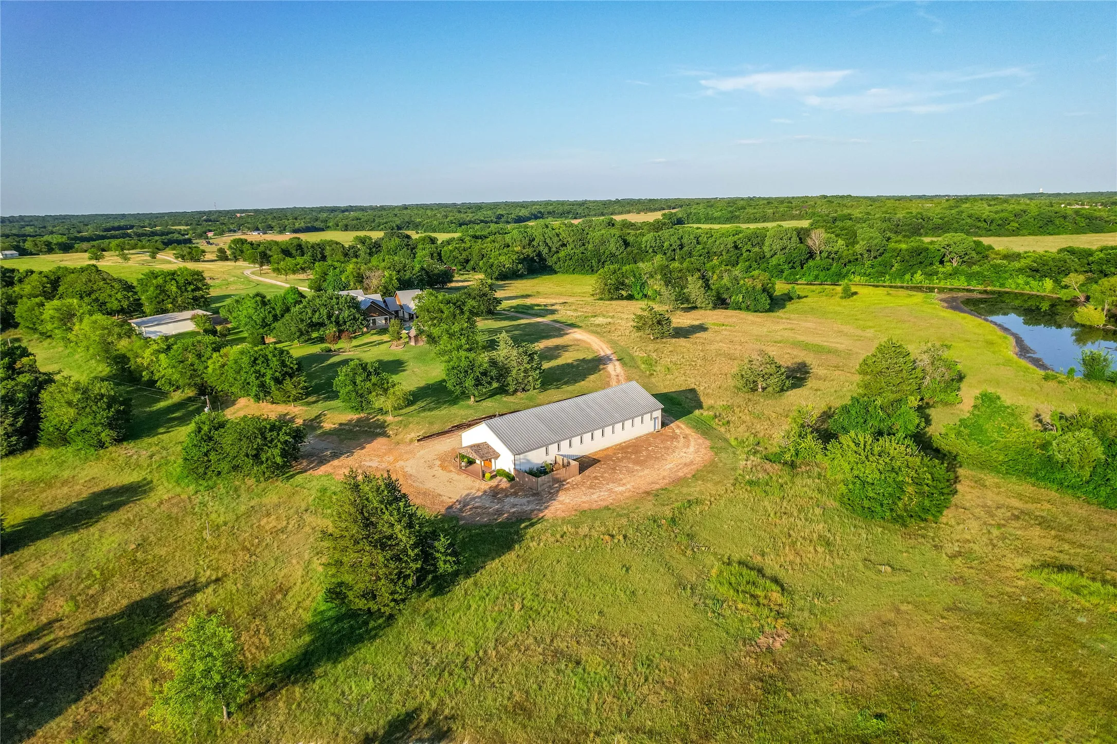 Expansive rural property featuring a large outbuilding with multiple windows, a winding dirt road, scattered mature trees, and a natural pond