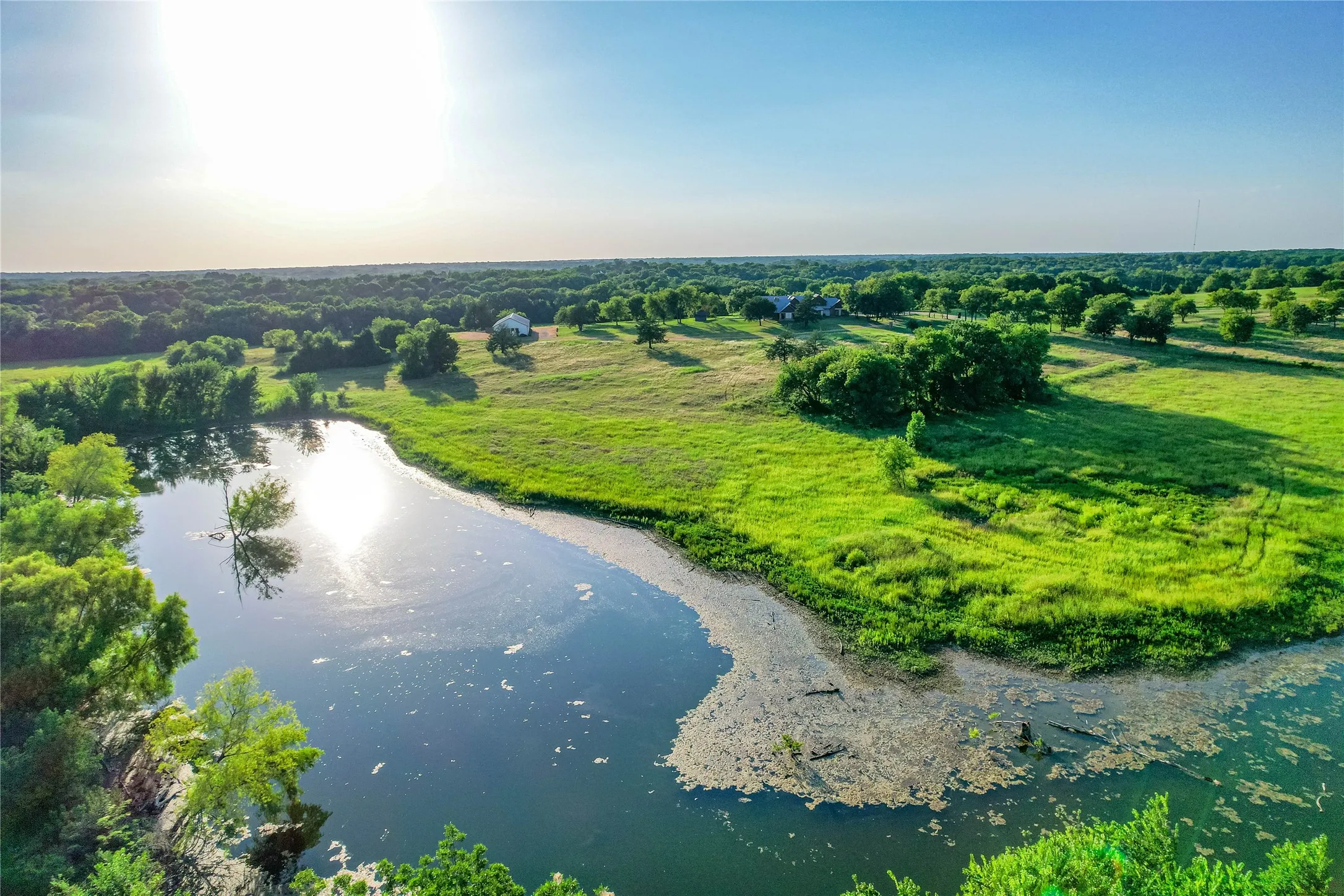Expansive natural landscape featuring a substantial body of water, lush green fields, and mature trees
