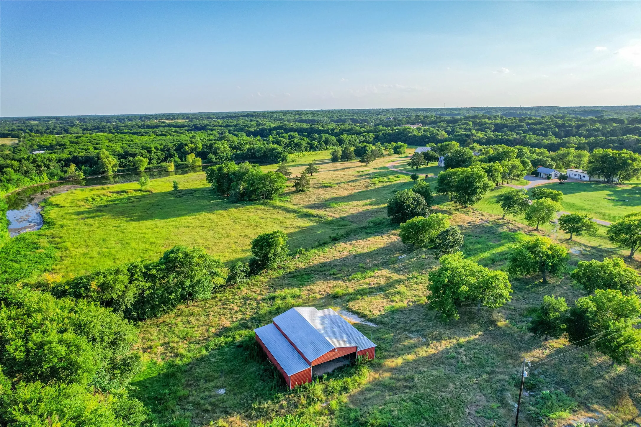 Expansive agricultural land featuring a red barn with a metal roof, mature trees, and a winding creek
