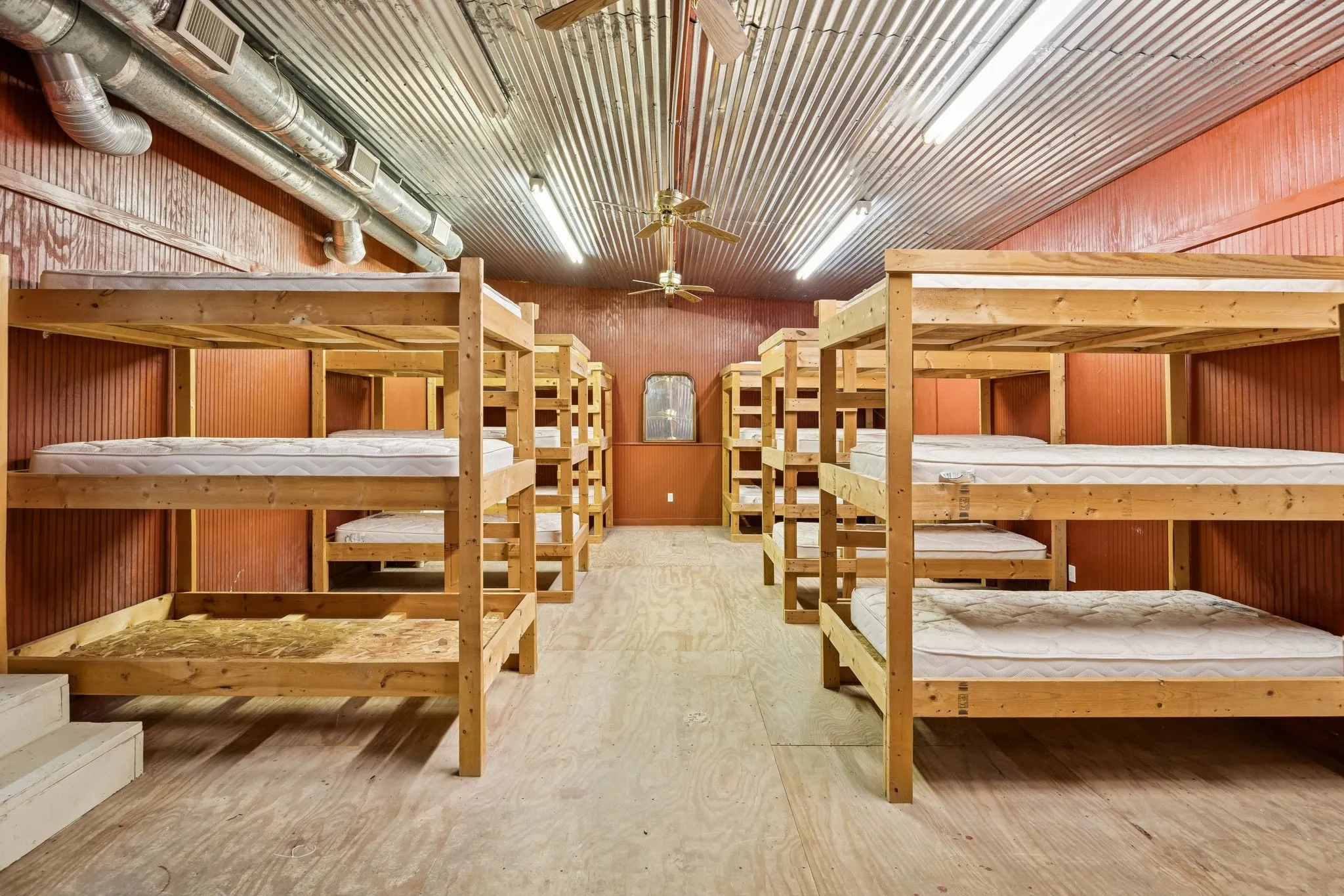 Accommodations room featuring multiple wooden bunk bed frames, corrugated metal ceiling, exposed ductwork, and fluorescent lighting