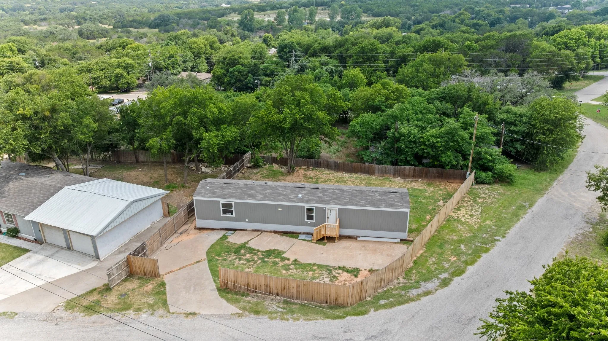 Gray residential structure with a light gray roof, featuring a wooden entry deck, perimeter fencing, and a concrete driveway