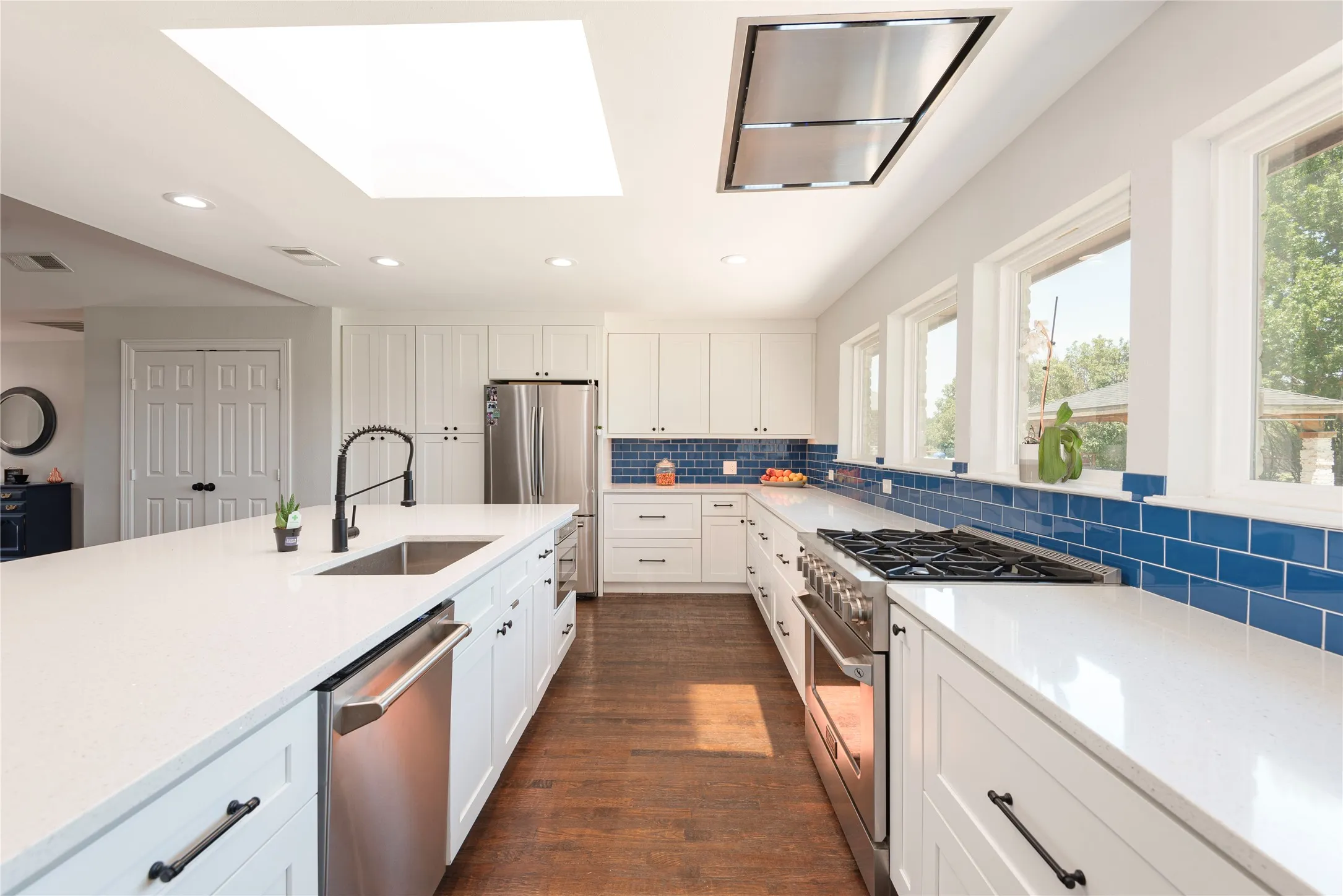 Bright kitchen featuring white cabinetry, recessed vent, a large center island with integrated sink, stainless steel appliances, a six-burner propane gas range, and a vibrant blue tile backsplash
