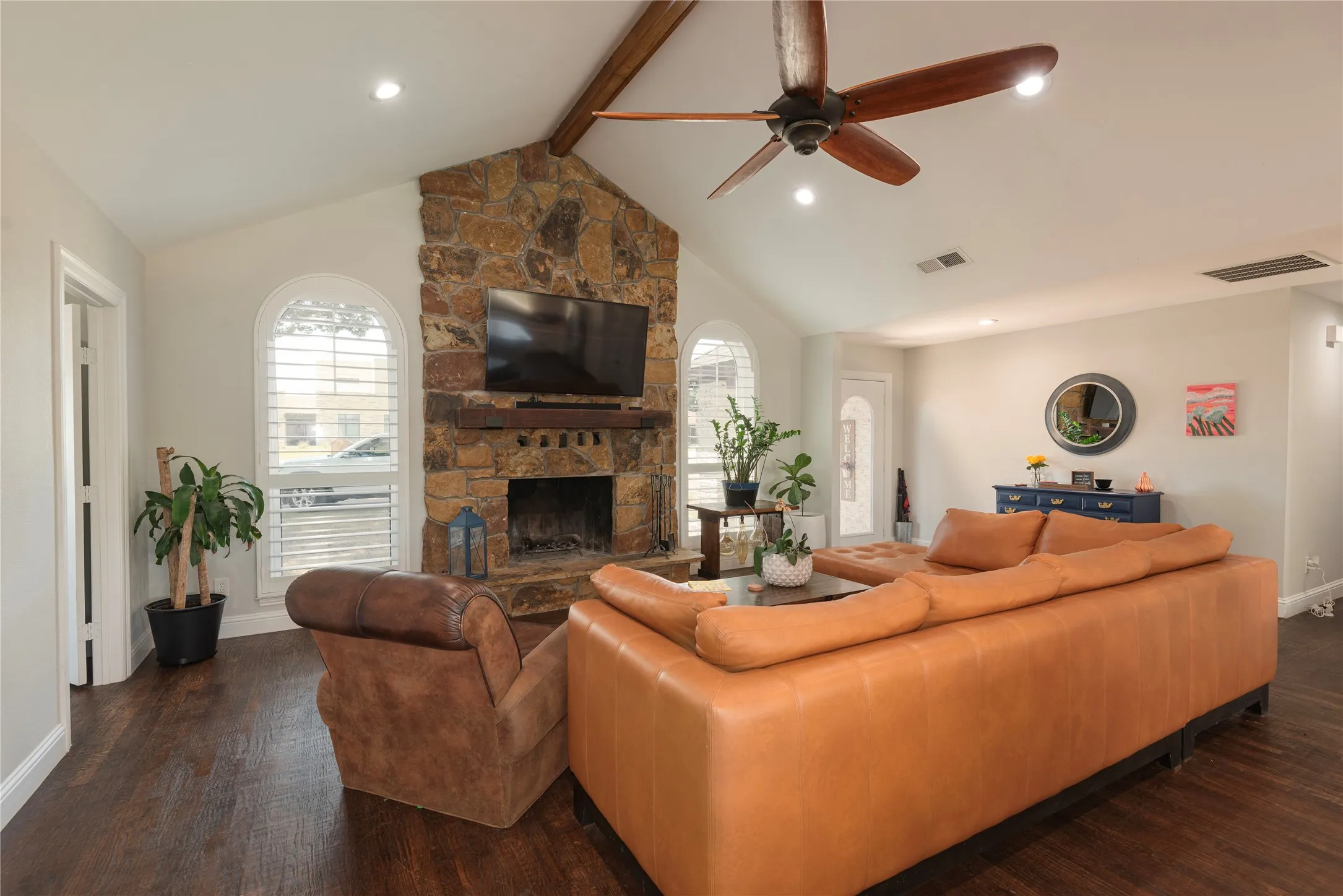 Vaulted ceiling living area featuring a stone-faced fireplace with a wood mantel, dark wood-finish flooring, and arched windows with plantation shutters