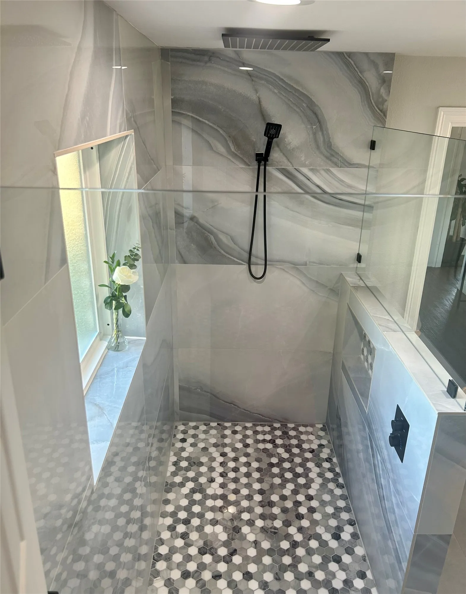 Oversized walk-in shower featuring large wall tiles, a built-in window, a rain shower head, handheld sprayer, hexagonal mosaic floor tiles, rainfall showerhead and wand, out of sight large niche for oversized bottles