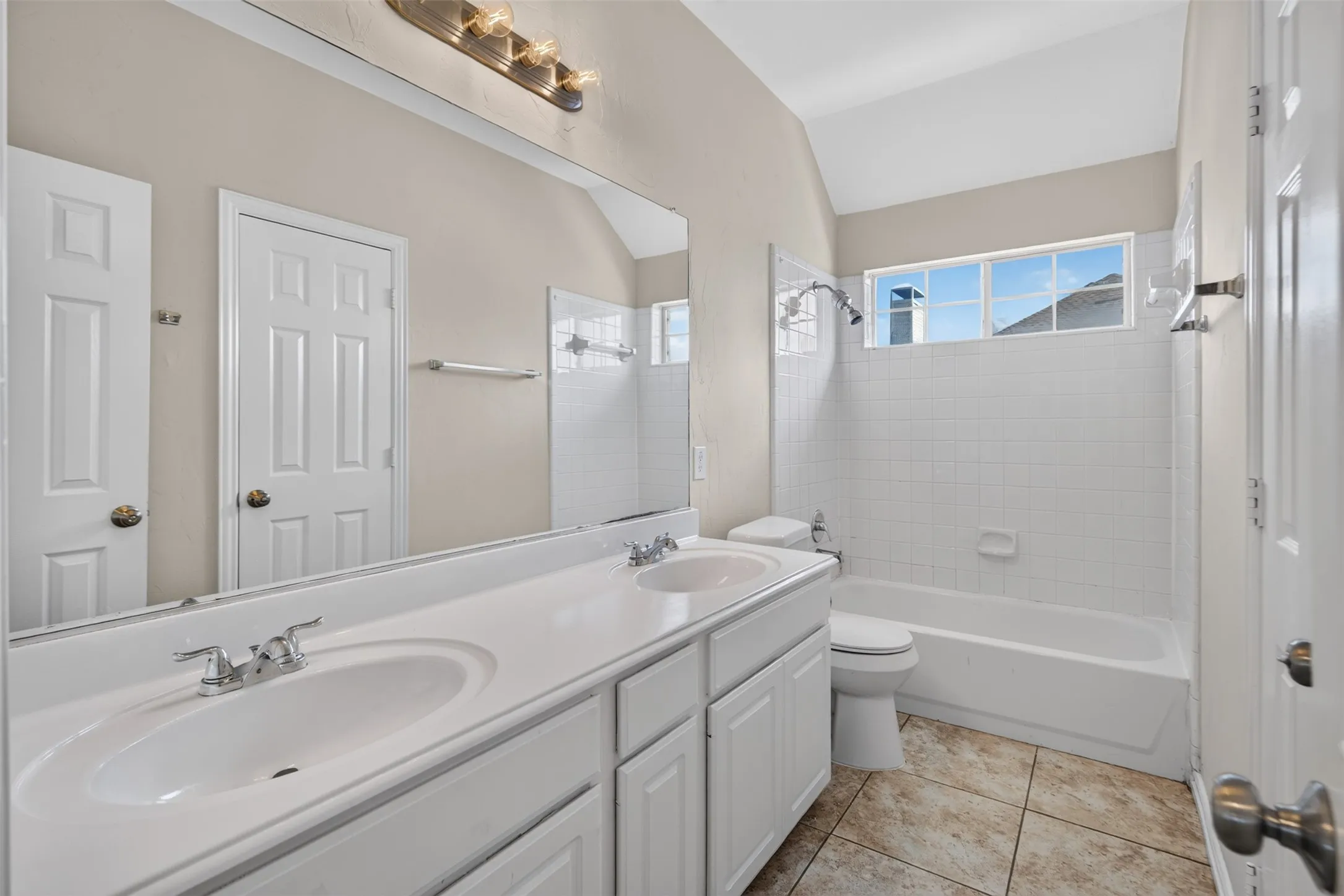 Double vanity with white cabinetry and integrated sinks, large wall-mounted mirror, ceramic tile flooring, bathtub with shower, and an arched ceiling
