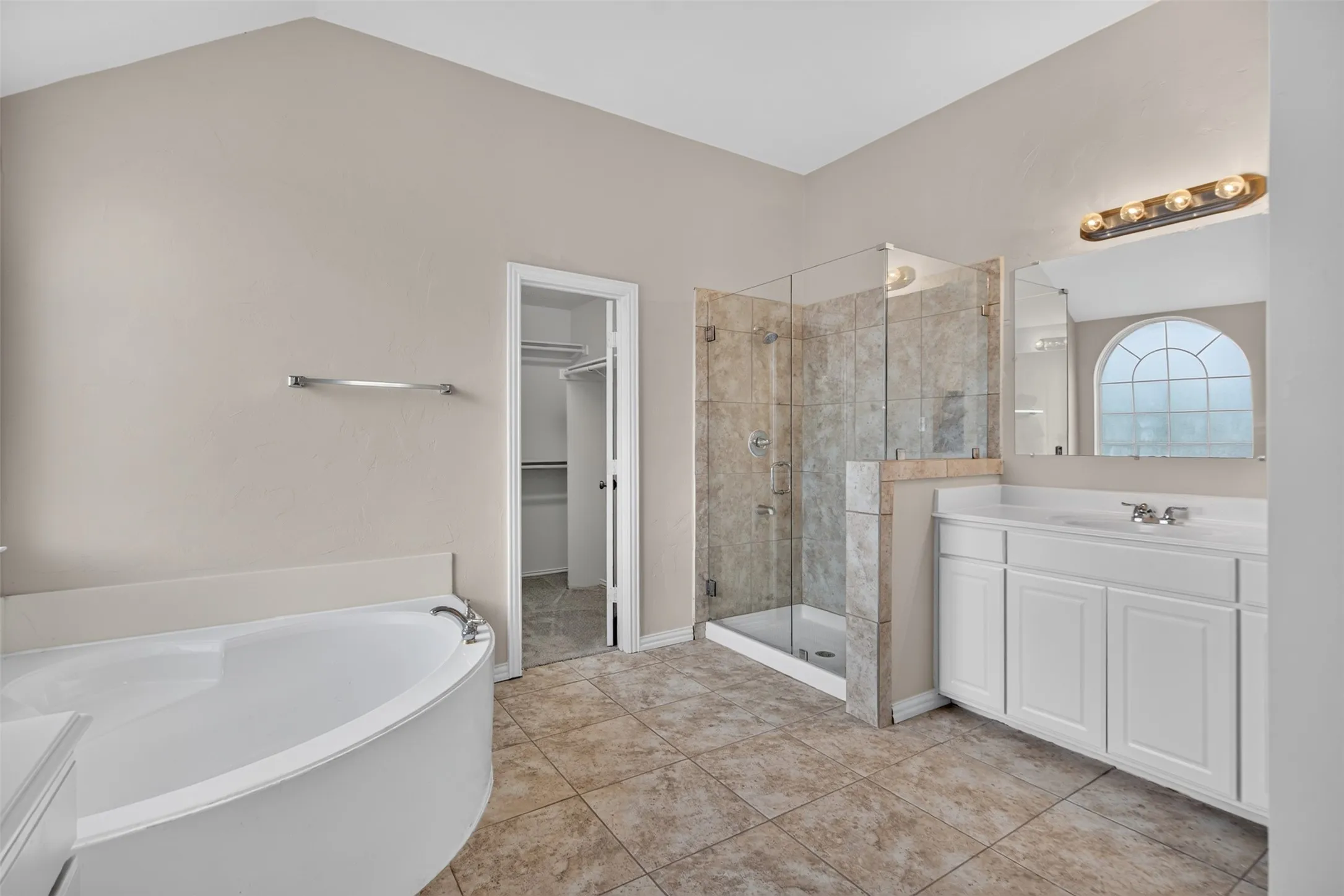 Bathroom featuring a corner soaking tub, a glass-enclosed shower with tiled walls, a single vanity with storage, a large mirror, and tiled flooring
