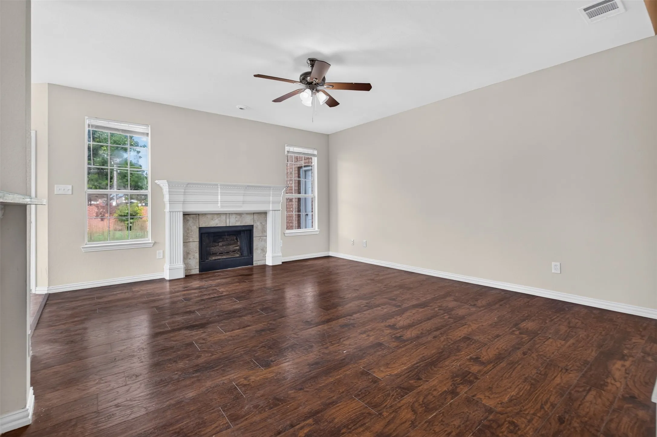 Spacious room featuring wood-finish flooring, a decorative fireplace with a tile surround and substantial mantel, multiple windows, and a ceiling fan