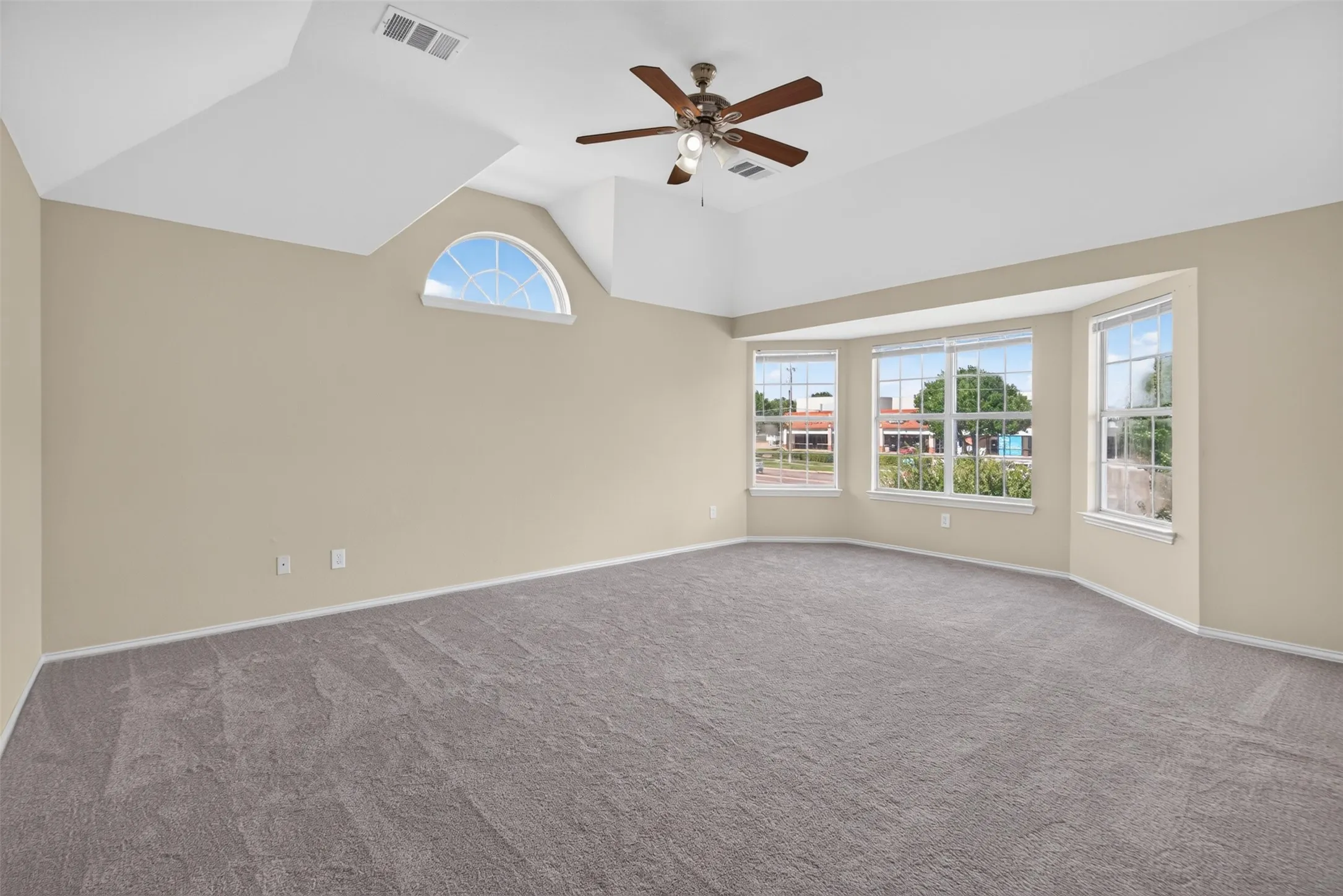Spacious room featuring vaulted ceilings, a ceiling fan, bay windows, an arched transom window, and neutral-toned carpeting