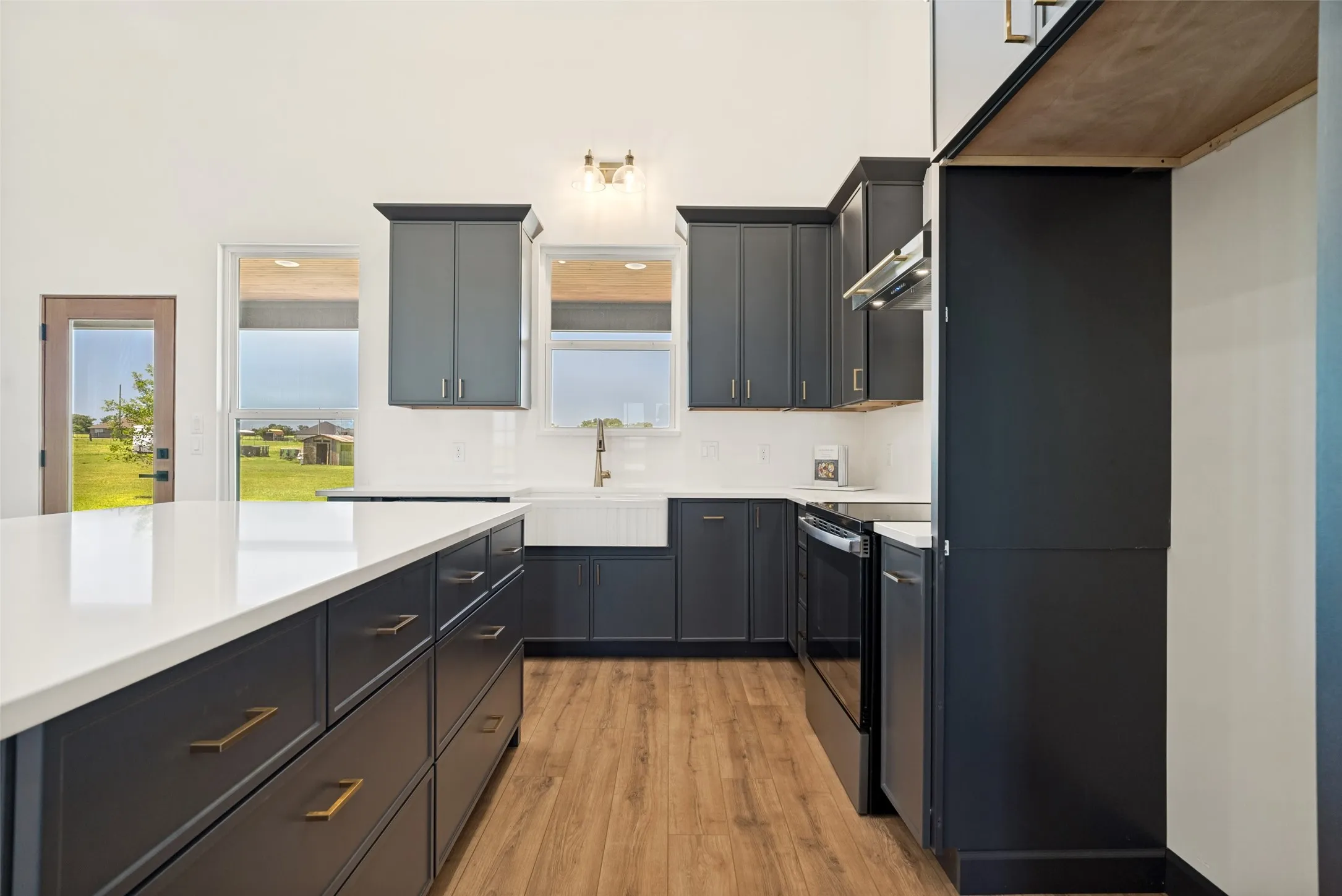 Modern kitchen featuring dark cabinetry, white countertops, wood-finish flooring, a farmhouse sink, and stainless steel appliances