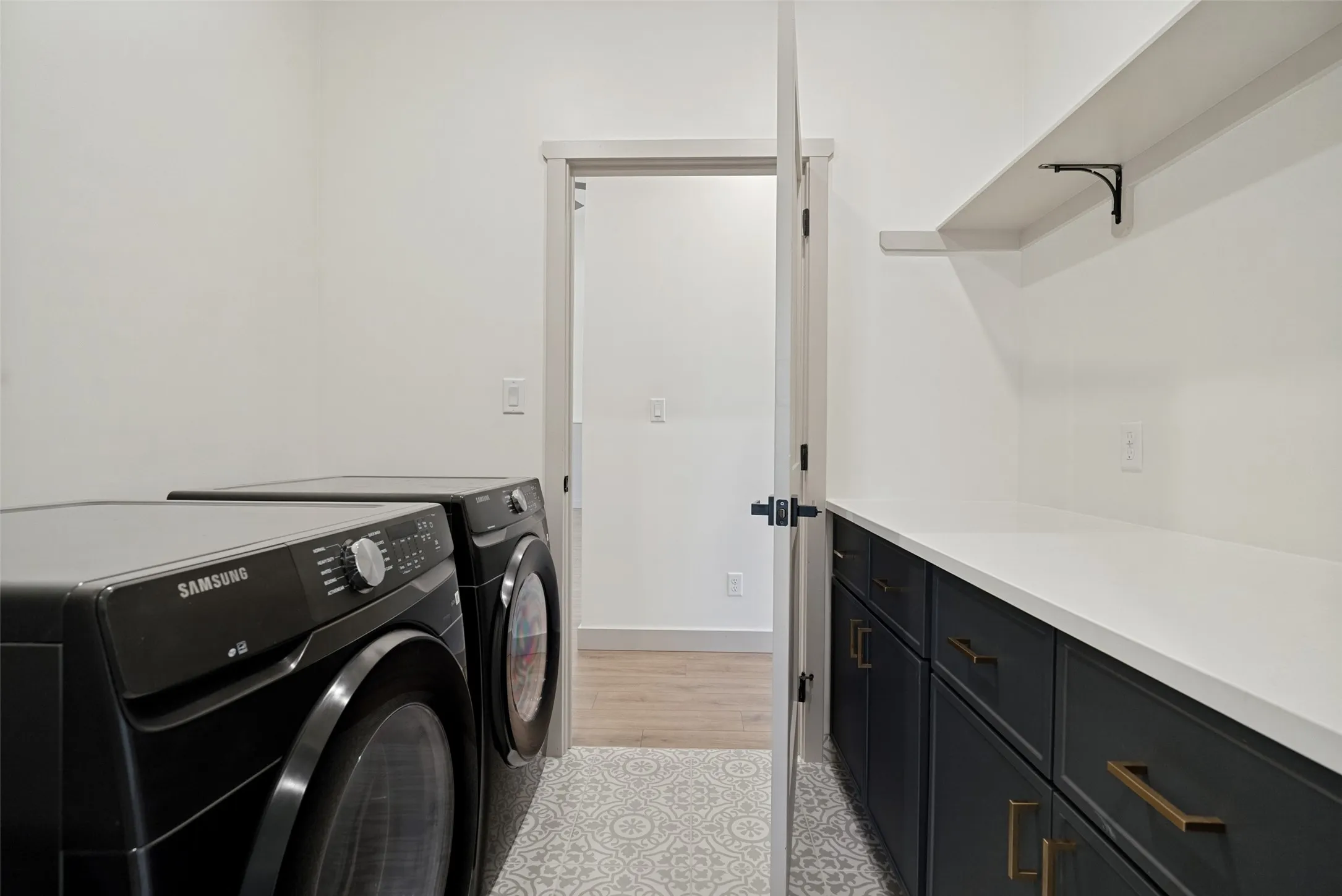 Laundry area featuring patterned tile flooring, dark cabinetry with brass hardware, a white countertop, and an upper wall shelf
