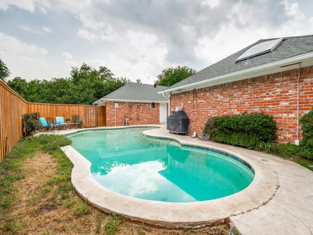 Curved swimming pool with concrete decking, surrounded by a wood privacy fence and brick exterior wall