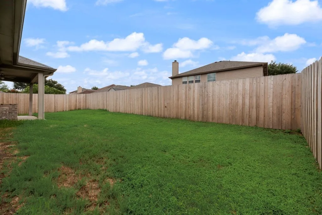 Expansive backyard featuring a lush green lawn, full wood privacy fence, and covered patio with a built-in stone feature