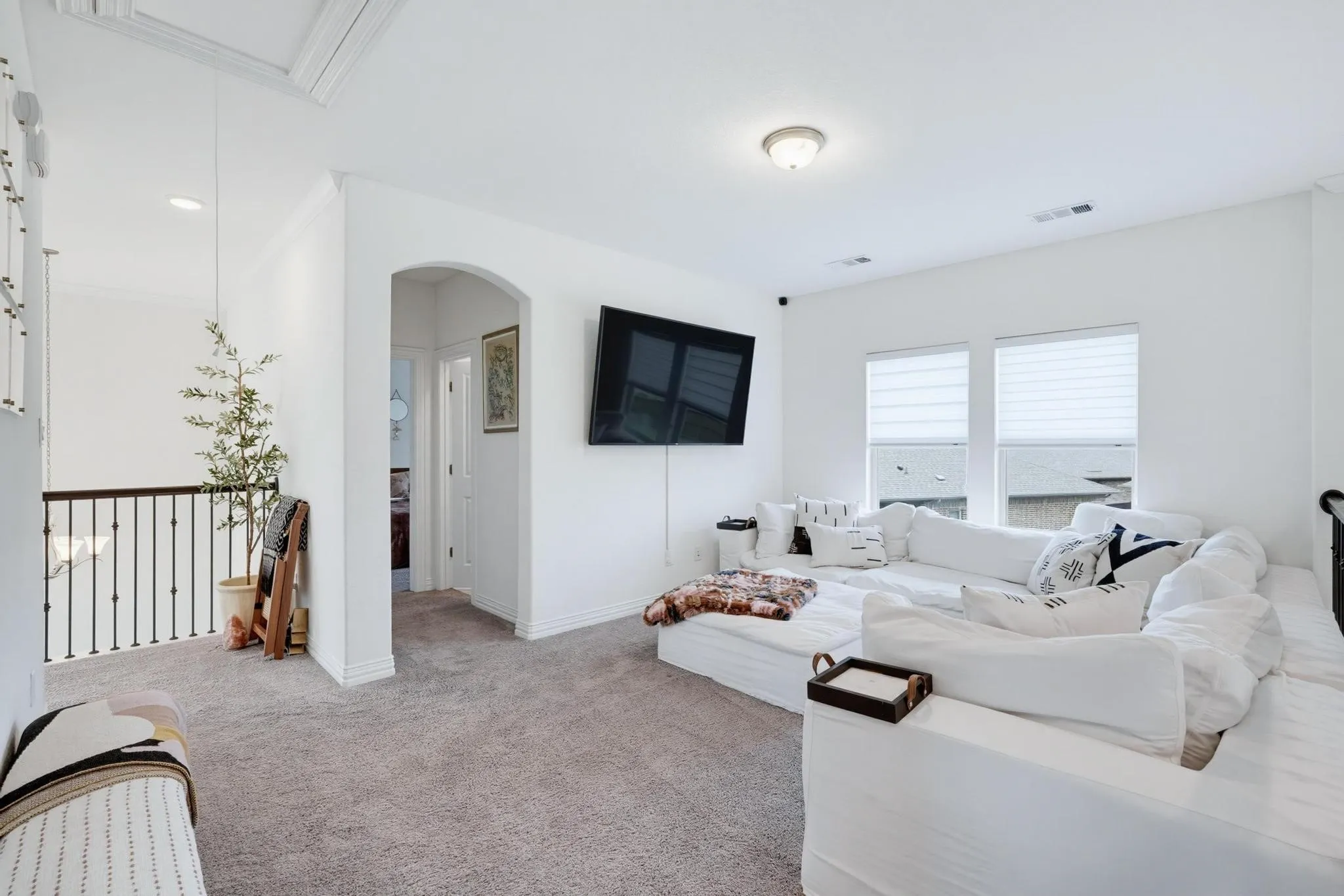 Carpeted open loft area featuring a wall-mounted display, an arched doorway, and two windows with blinds