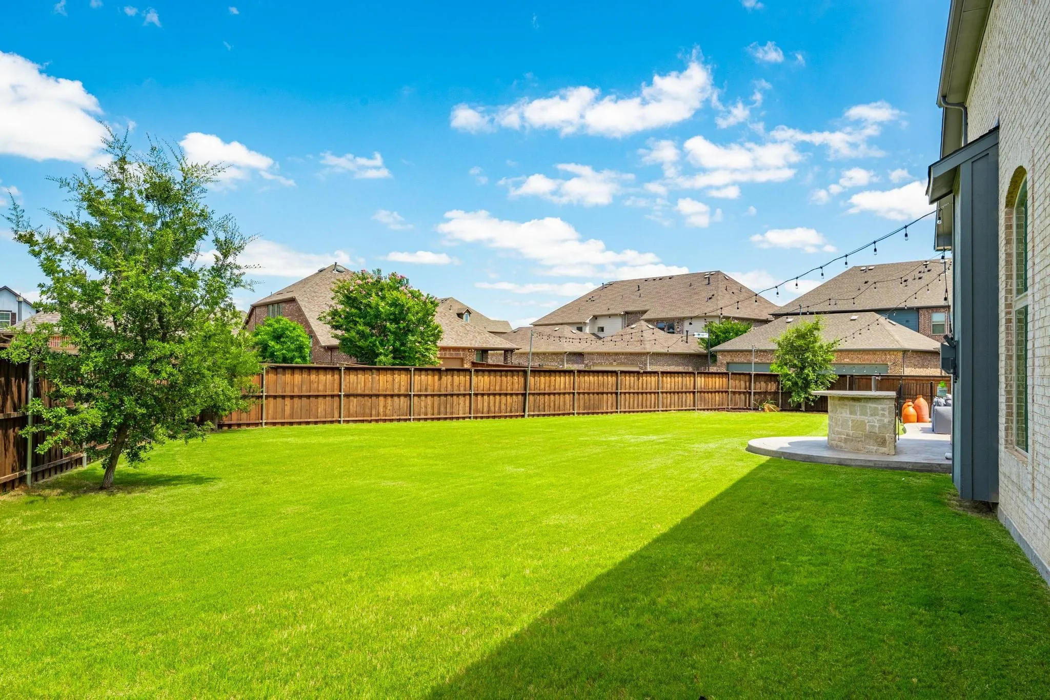 Expansive green lawn framed by a privacy fence, featuring a mature tree, stone patio, and string lighting