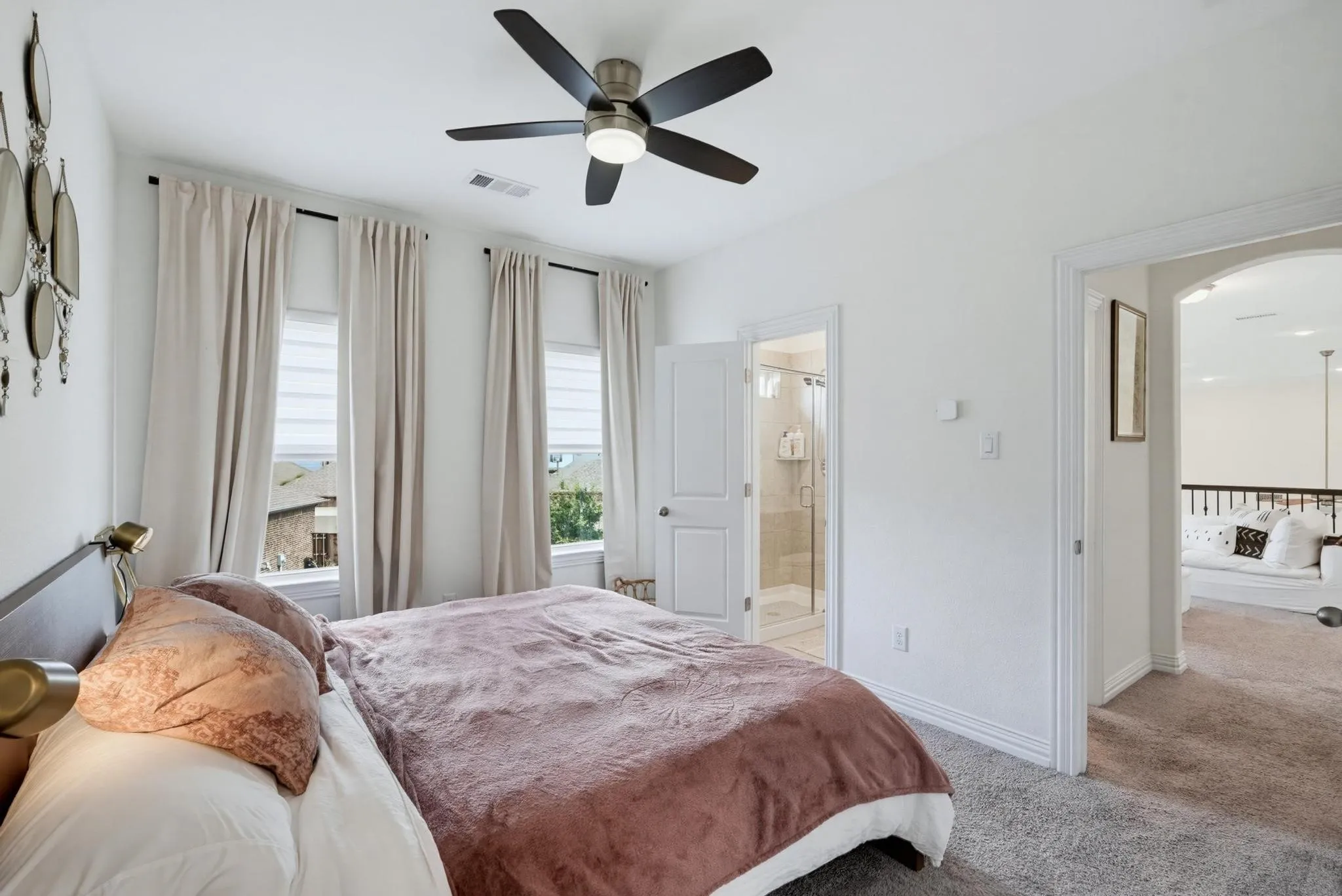 Carpeted bedroom featuring three windows with blinds and drapes, a ceiling fan with integrated lighting, an arched doorway, and a white paneled door revealing a tiled shower with a glass enclosure