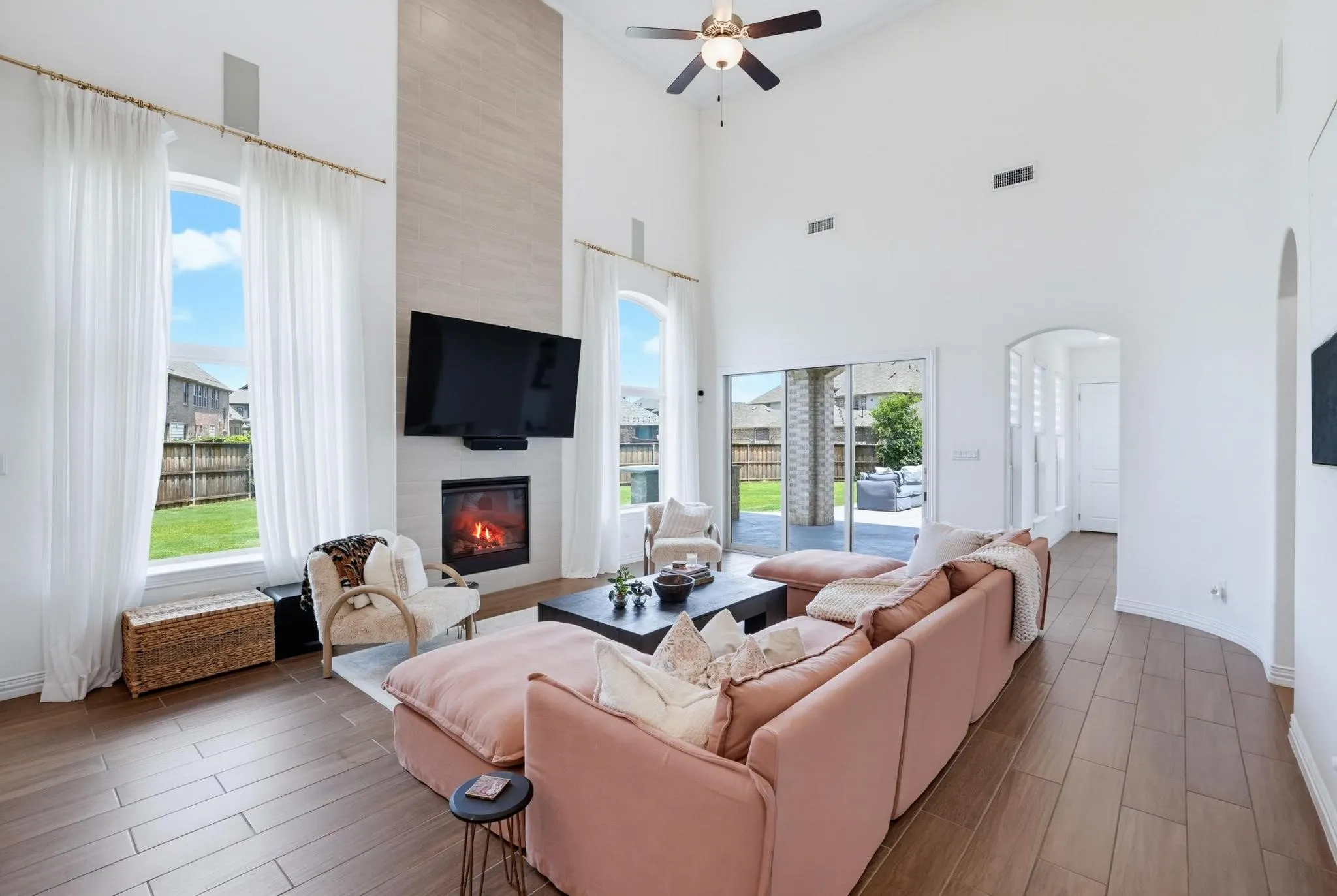 Spacious living area featuring a two-story ceiling, wood-finish flooring, a contemporary fireplace with a mounted television above, and large arched windows with sheer curtains