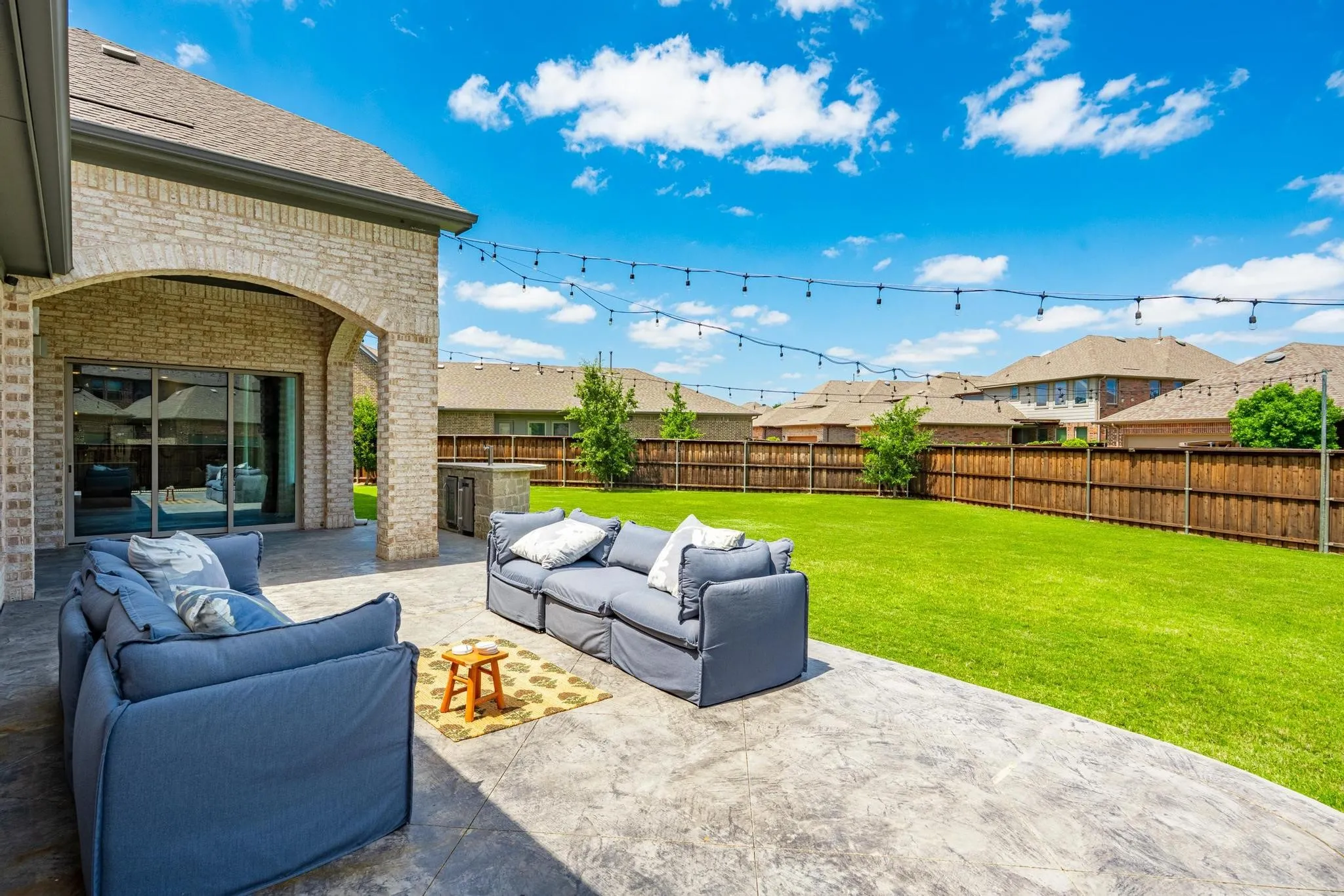 Spacious concrete patio featuring an arched brick-clad covered area, built-in outdoor kitchen with stone finish, and string lighting