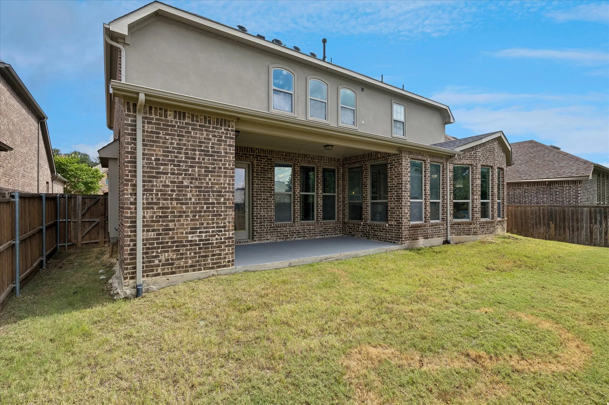Expansive backyard featuring a covered patio, brick and stucco exterior, multiple arched windows on the upper level, and a surrounding privacy fence
