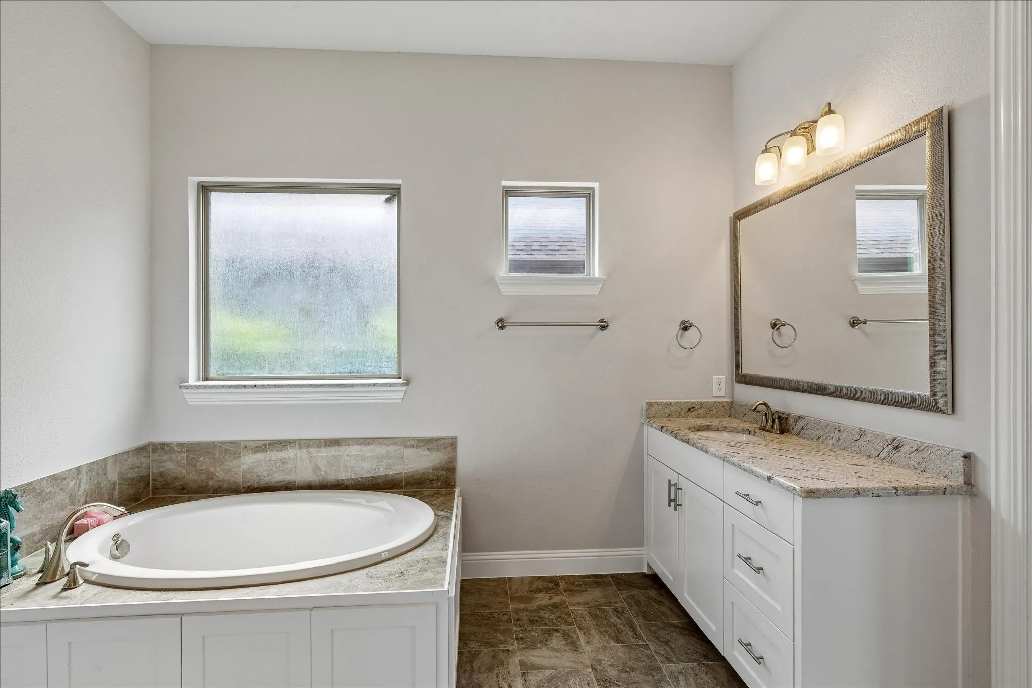 Bathroom featuring a built-in soaking tub with surrounding tile, a single vanity with stone countertop, and a large framed mirror