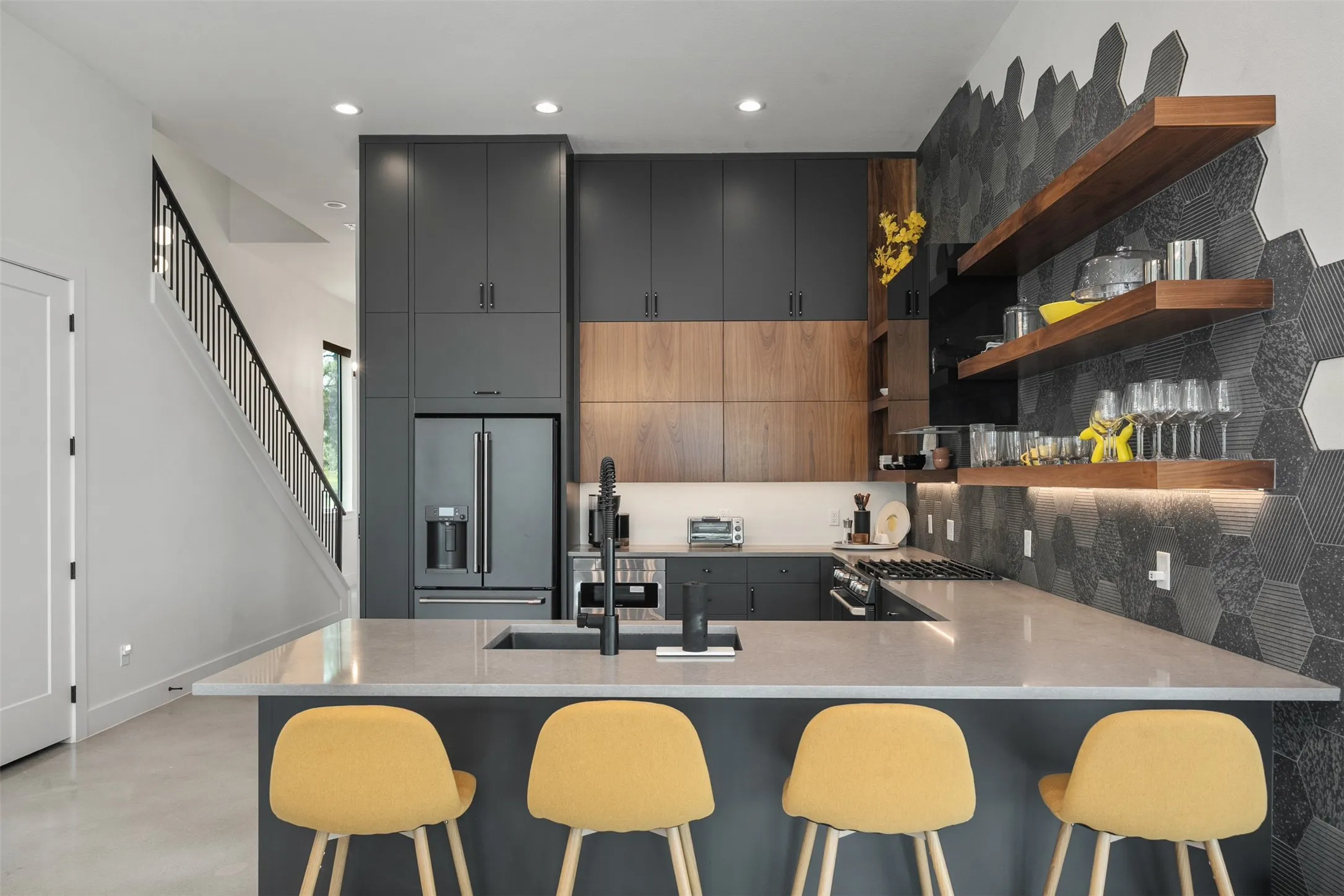 Modern kitchen featuring dark gray and wood-finish cabinetry, a large kitchen island with integrated sink, stainless steel appliances, and a unique hexagonal tile backsplash