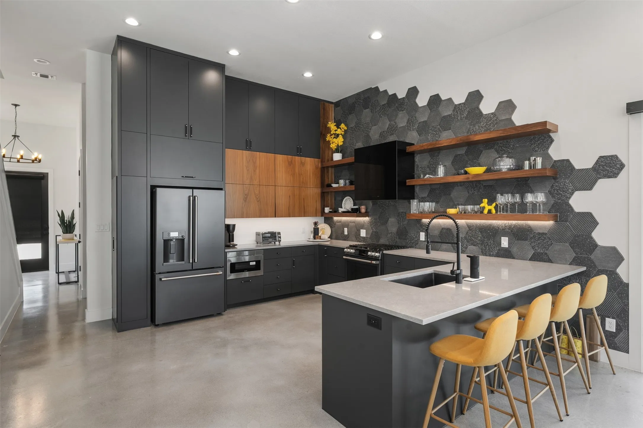 Modern kitchen featuring concrete flooring, dark flat-panel cabinetry, a hexagon tile accent wall, built-in stainless steel appliances, and a central island with an undermount sink and light-toned countertop