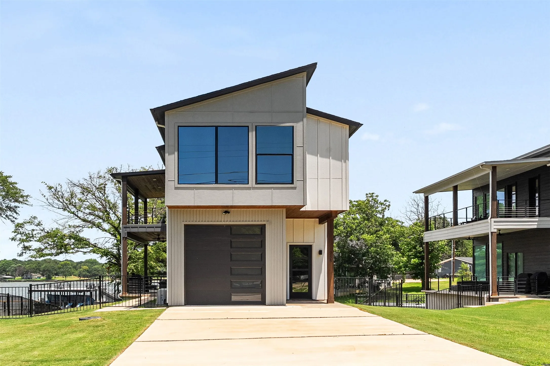 Contemporary waterfront residence featuring a modern dark-paneled garage door, multiple balconies, a front entry with vertical siding, a flat-pitched roof, and extensive perimeter fencing
