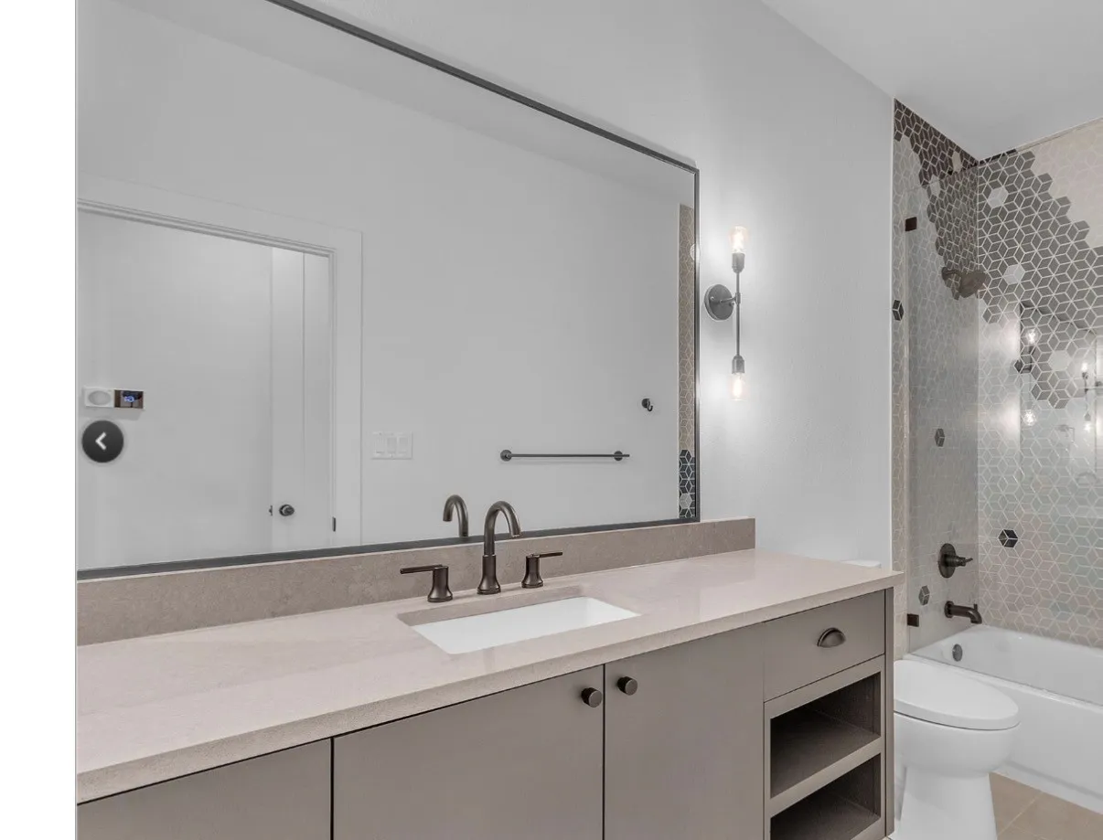 Contemporary bathroom featuring a spacious vanity with an integrated sink, a large wall-mounted mirror, modern wall sconces, a mosaic tiled shower surround, and a white bathtub