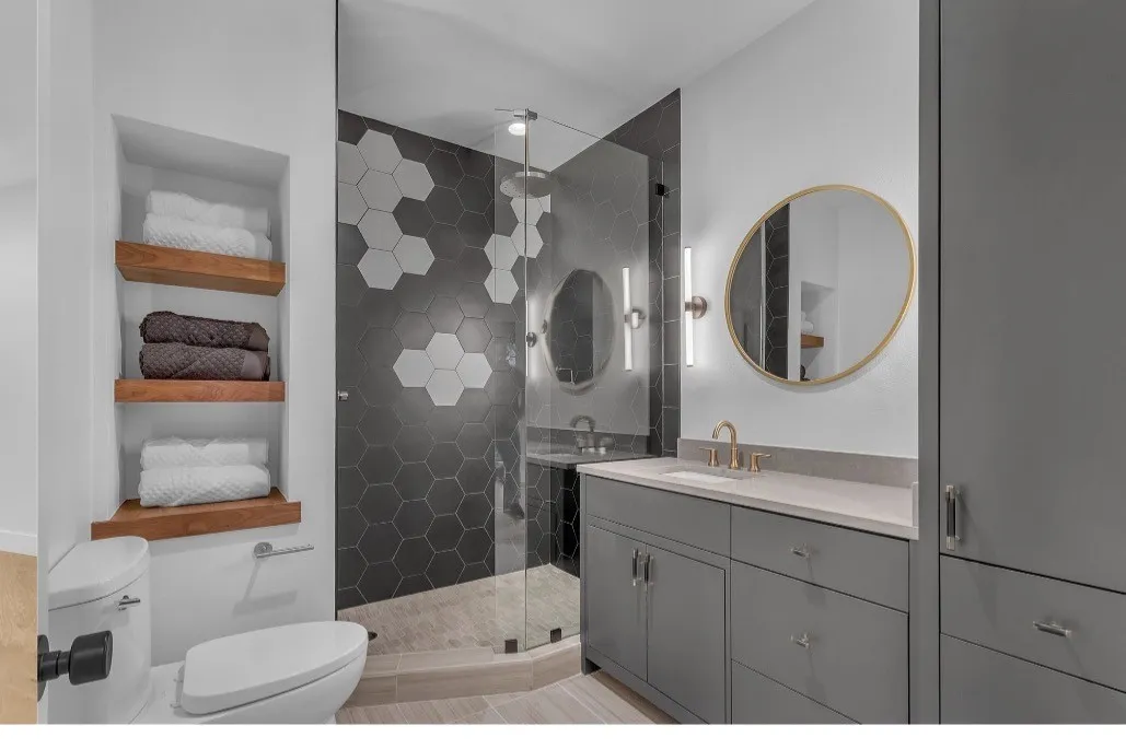 Bathroom featuring a frameless glass shower enclosure with hex tile surround, a single vanity with stone countertop, built-in storage cabinetry, a wall-mounted mirror, and recessed wood shelving