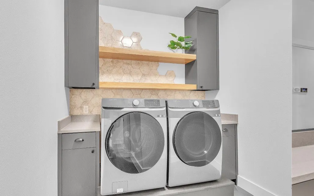 Dedicated laundry area featuring modern gray cabinetry, two wood-finish floating shelves, and a hexagonal tiled accent wall