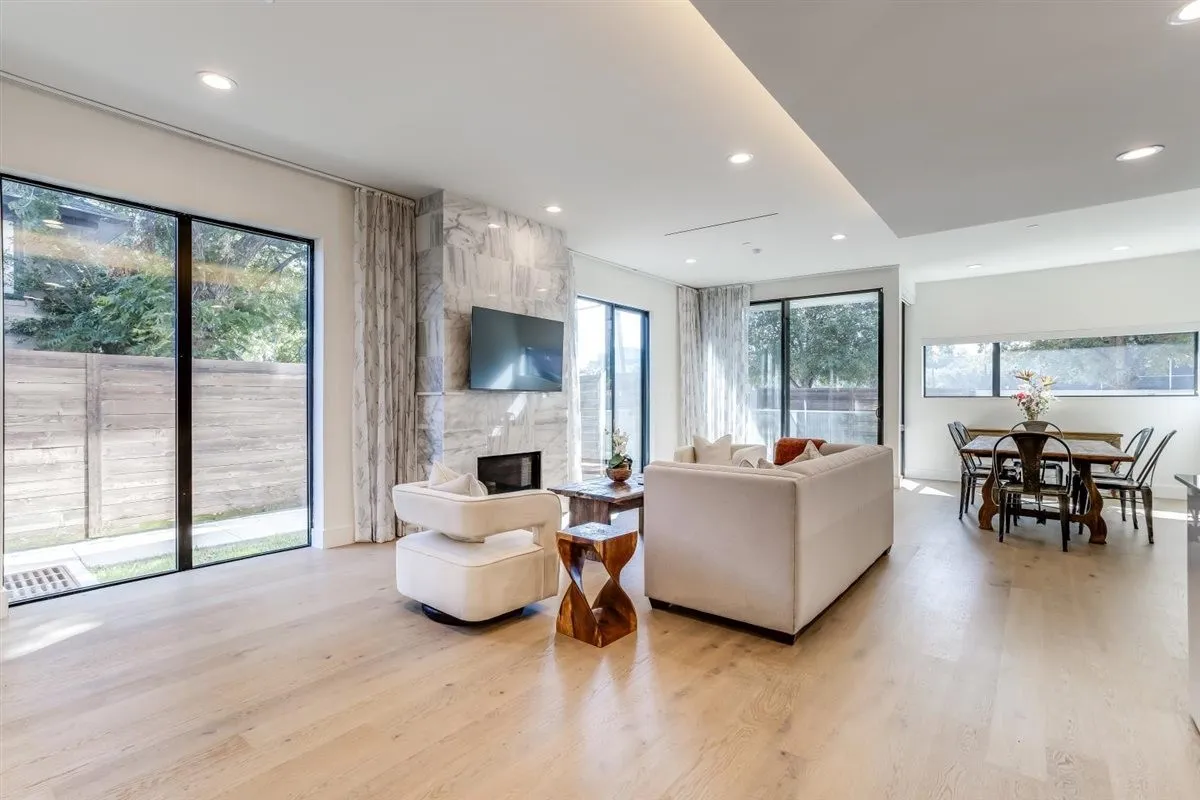 Open-concept living area featuring light wood-finish flooring, a contemporary fireplace with a stone surround, recessed lighting, and large windows with black frames